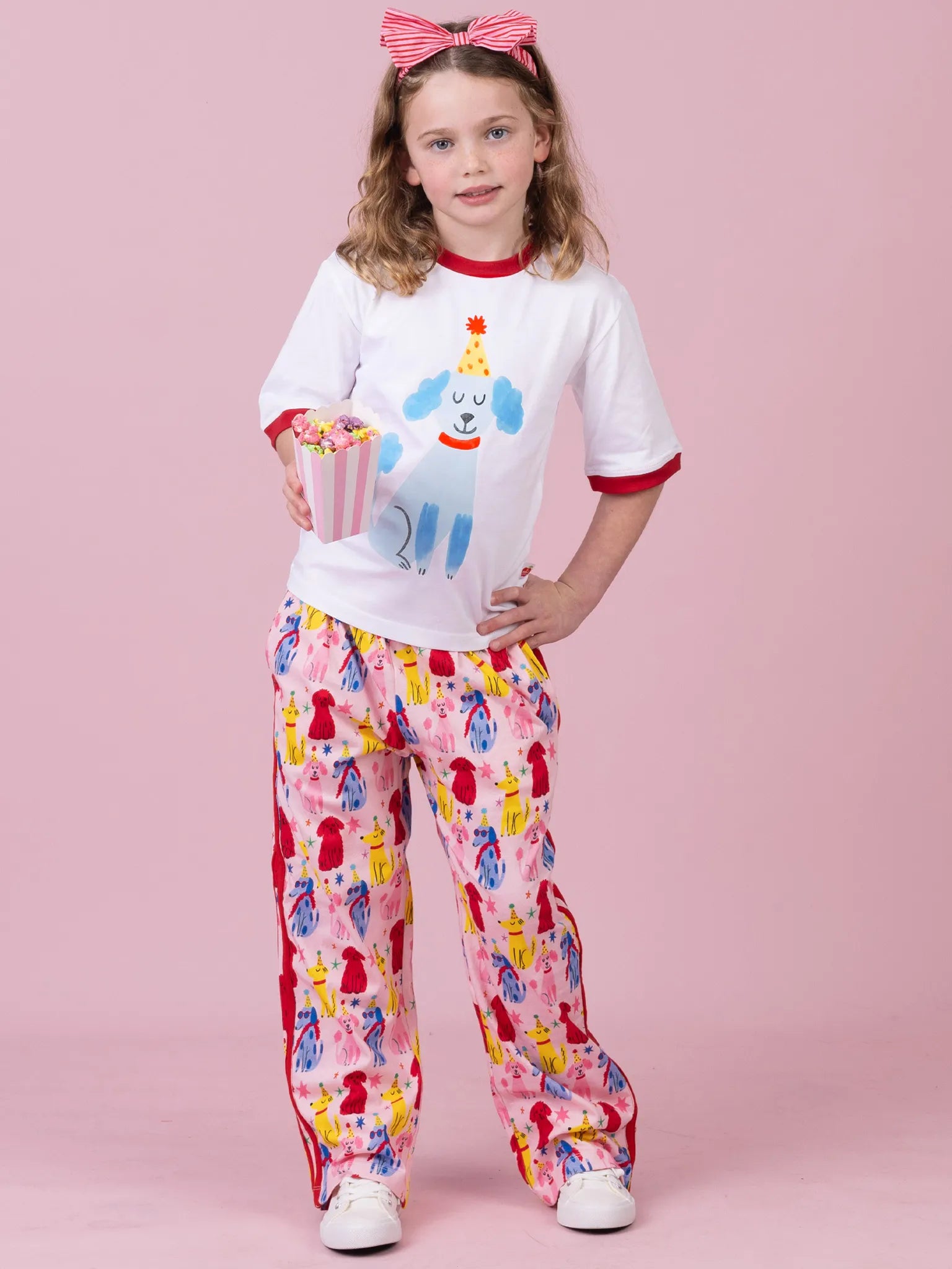 A young girl modeling a white t-shirt with a blue cartoon dog graphic and red trim, paired with pink pants featuring a colorful dog party pattern, standing against a solid pink background.