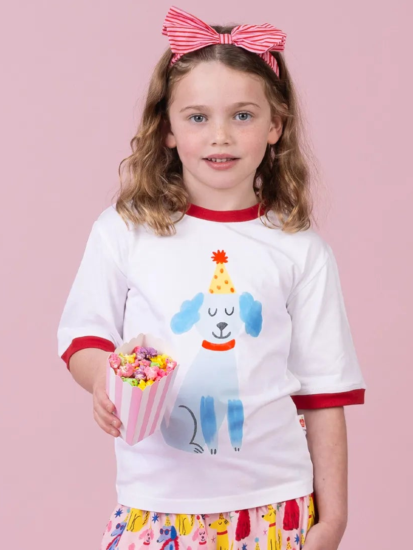 A young girl with a pink and white striped bow in her hair wears a white ringer T-shirt with red trim and a cartoon dog graphic, holding a striped popcorn bucket against a solid pink background.