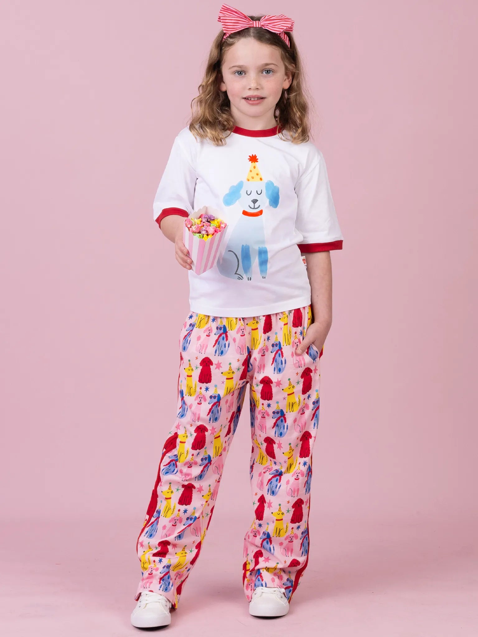 A young girl stands against a solid pink studio background wearing a white graphic t-shirt with red trim and a blue dog illustration, paired with pink pants covered in a colorful red, yellow, and blue pattern. She is holding a cup of popcorn and wearing a pink bow in her hair.