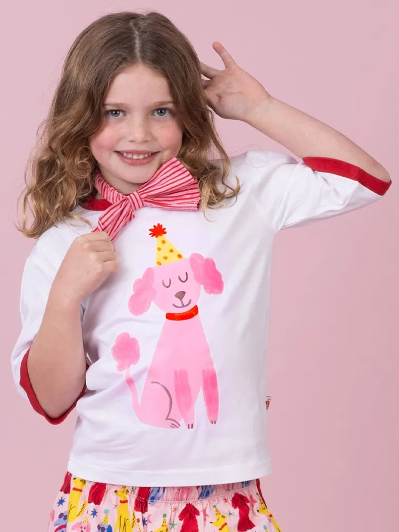A young girl with wavy light brown hair smiling and posing in a white short-sleeve t-shirt with red trim, featuring a pink poodle graphic wearing a party hat. She is also wearing a pink and white striped bow tie and a colorful patterned skirt, set against a solid light pink background.
