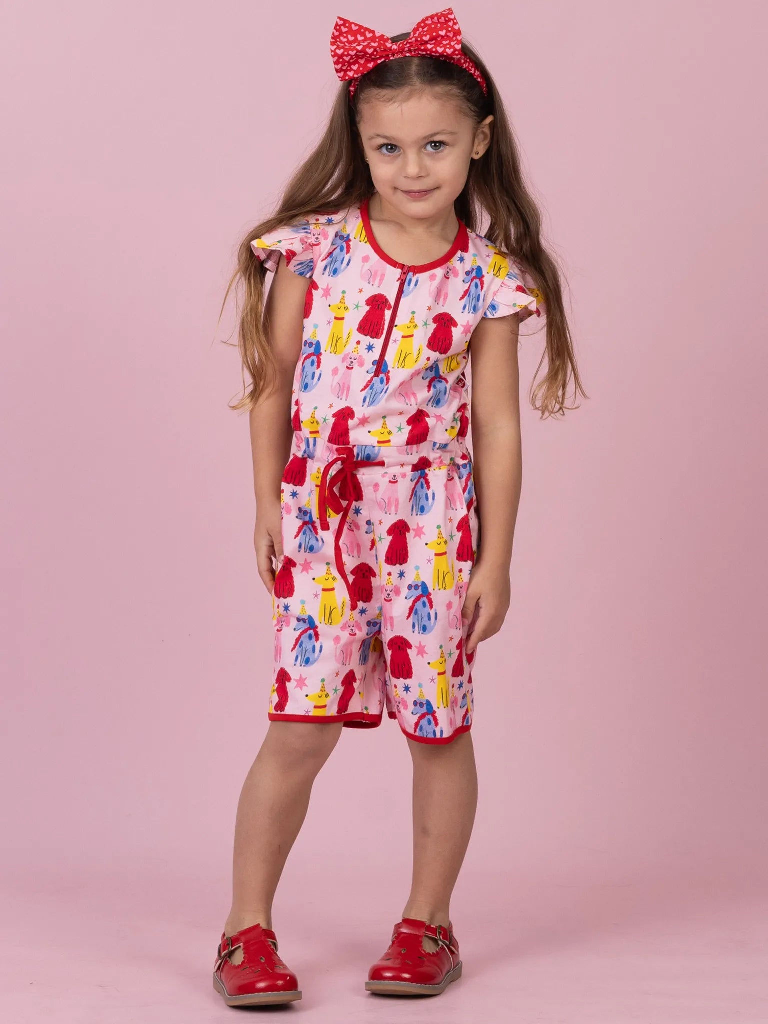 A young girl modeling a pink short-sleeve romper with a colorful cartoon dog print, a red drawstring waist, and red trim, paired with a red polka-dot bow headband and red shoes, against a solid pink background.