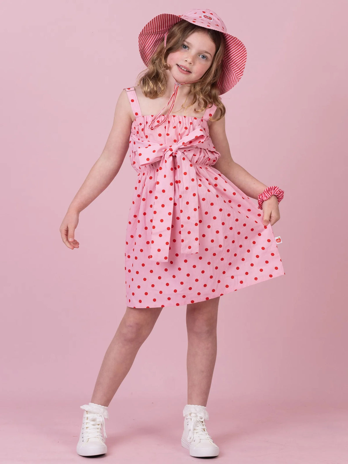 A young girl modeling a pink sleeveless dress with red polka dots, a matching pink sun hat, and white sneakers, posed against a solid pink studio background.