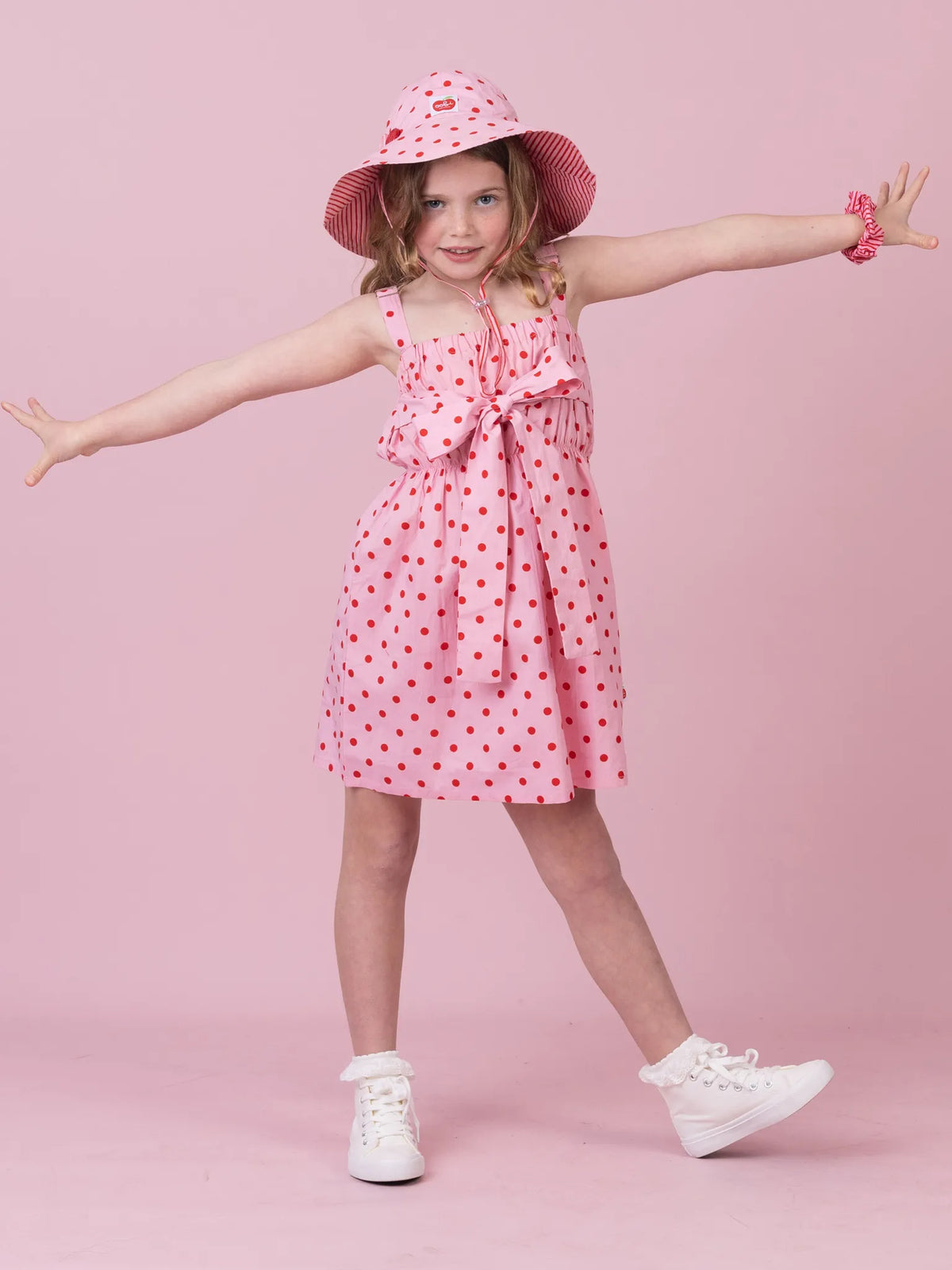 A young girl posing in a pink sleeveless dress with red polka dots, a matching pink sun hat with red dots, and white sneakers against a solid pink background.