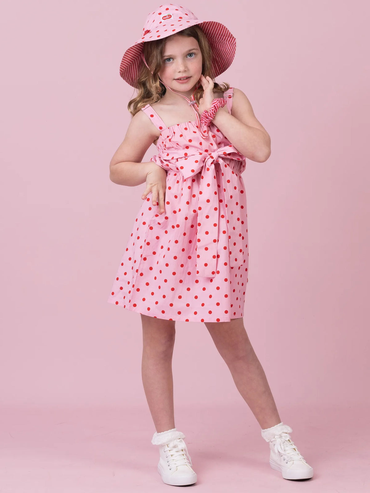 A young girl modeling a pink sleeveless dress with red polka dots, a matching pink sun hat, and white sneakers against a solid pink studio background.