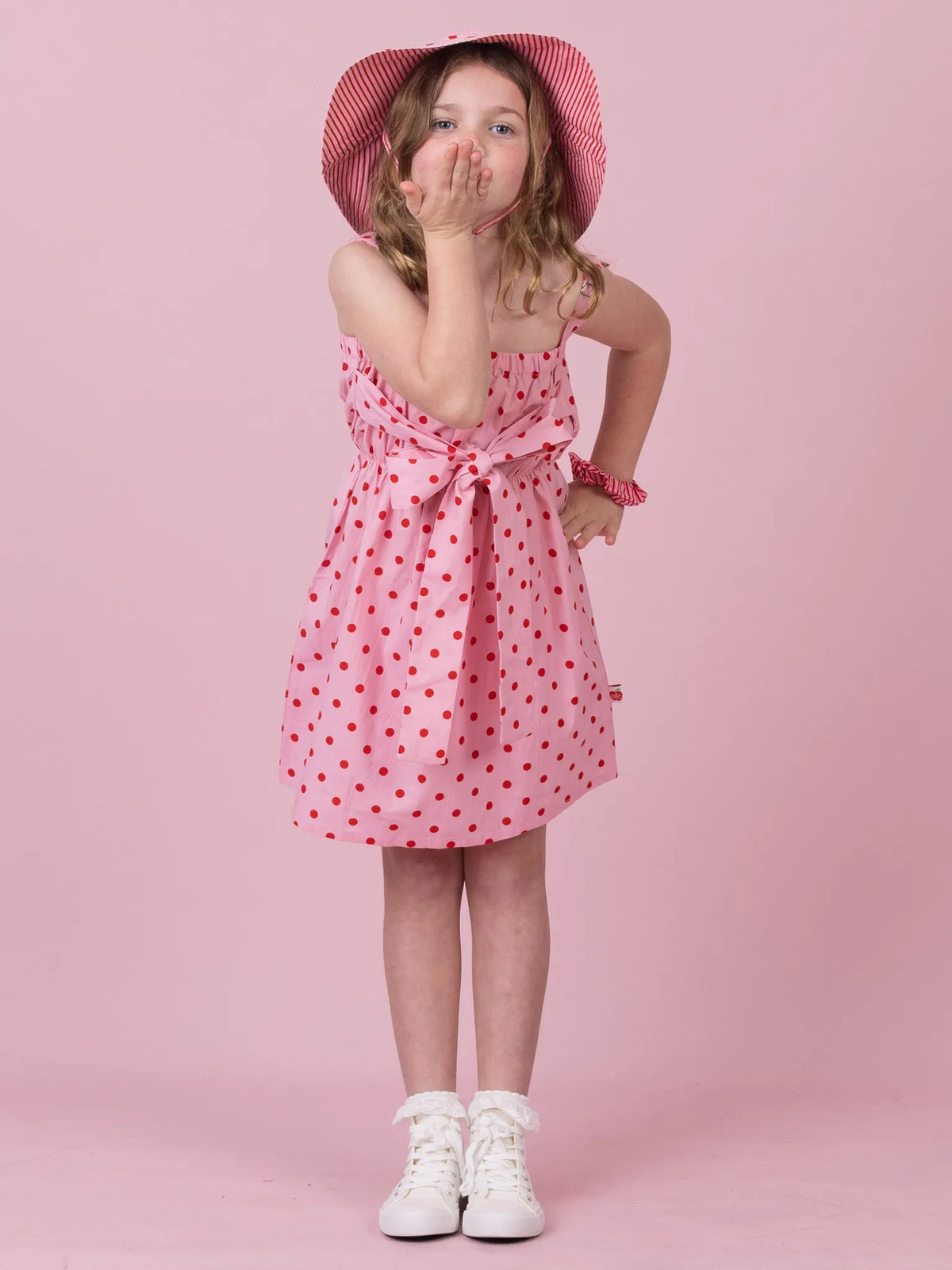 A young girl wearing a pink and red polka dot dress with a matching pink hat, posing against a solid pink studio background.