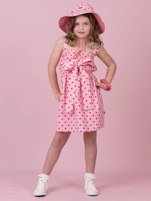 A young girl modeling a pink sleeveless dress with red polka dots, a matching pink sun hat, and white sneakers against a solid pink studio background.
