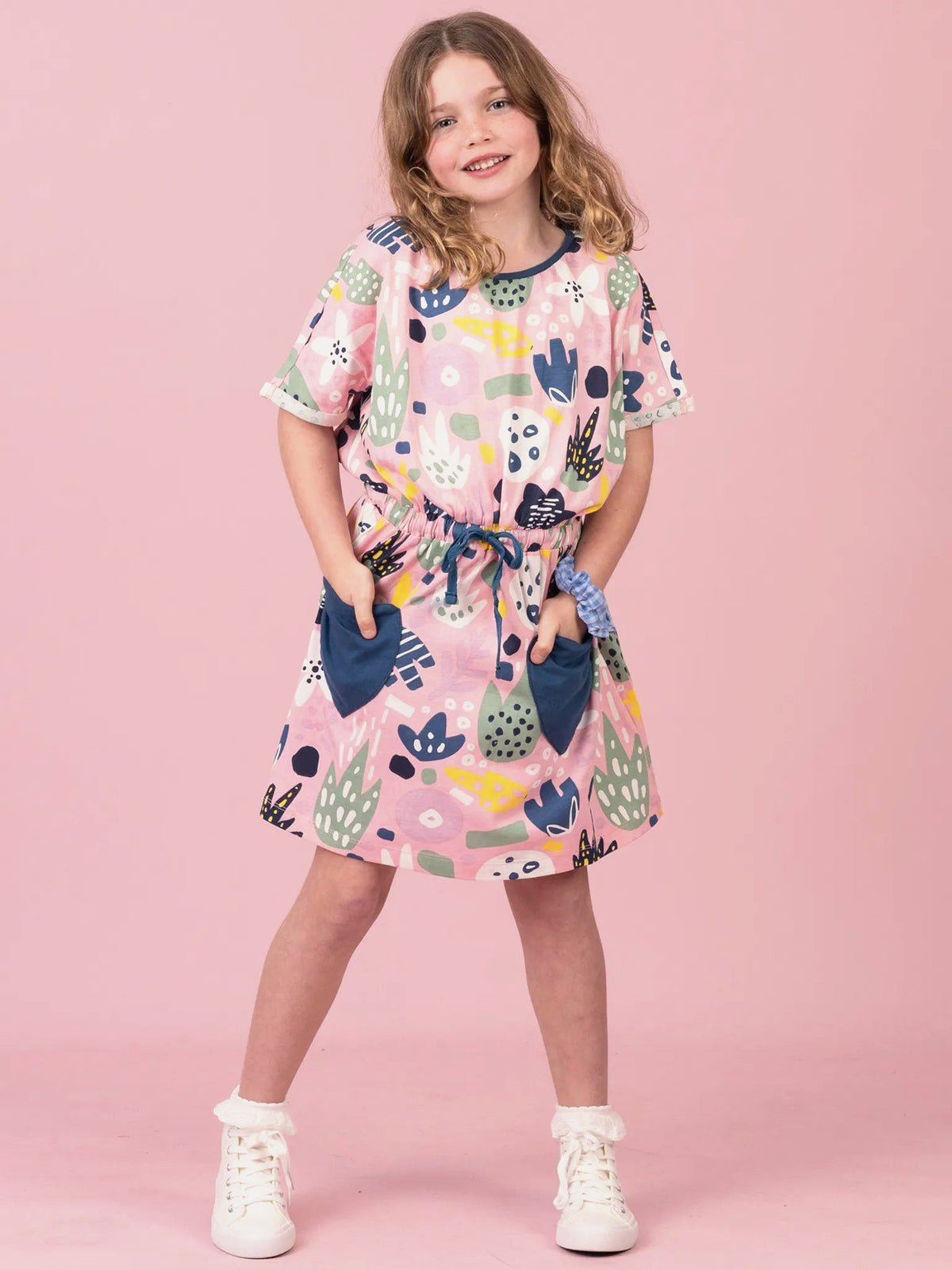 A young girl modeling a short-sleeve pink dress with a colorful abstract print, pockets, and a tie waist, standing against a solid pink background.
