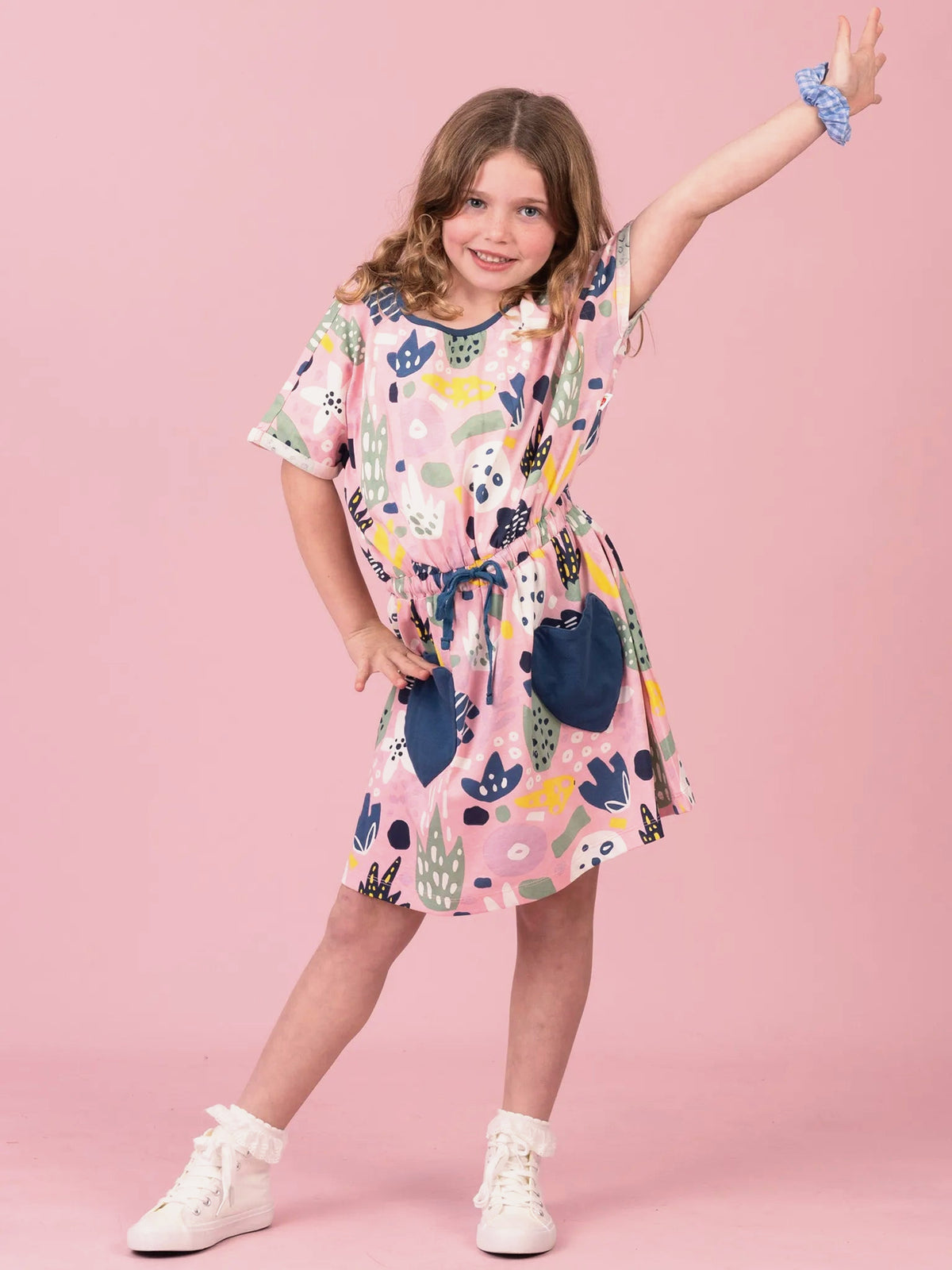 A young girl modeling a short-sleeve pink dress with a colorful abstract pattern and dark blue patch pockets, posed against a solid pink studio background.