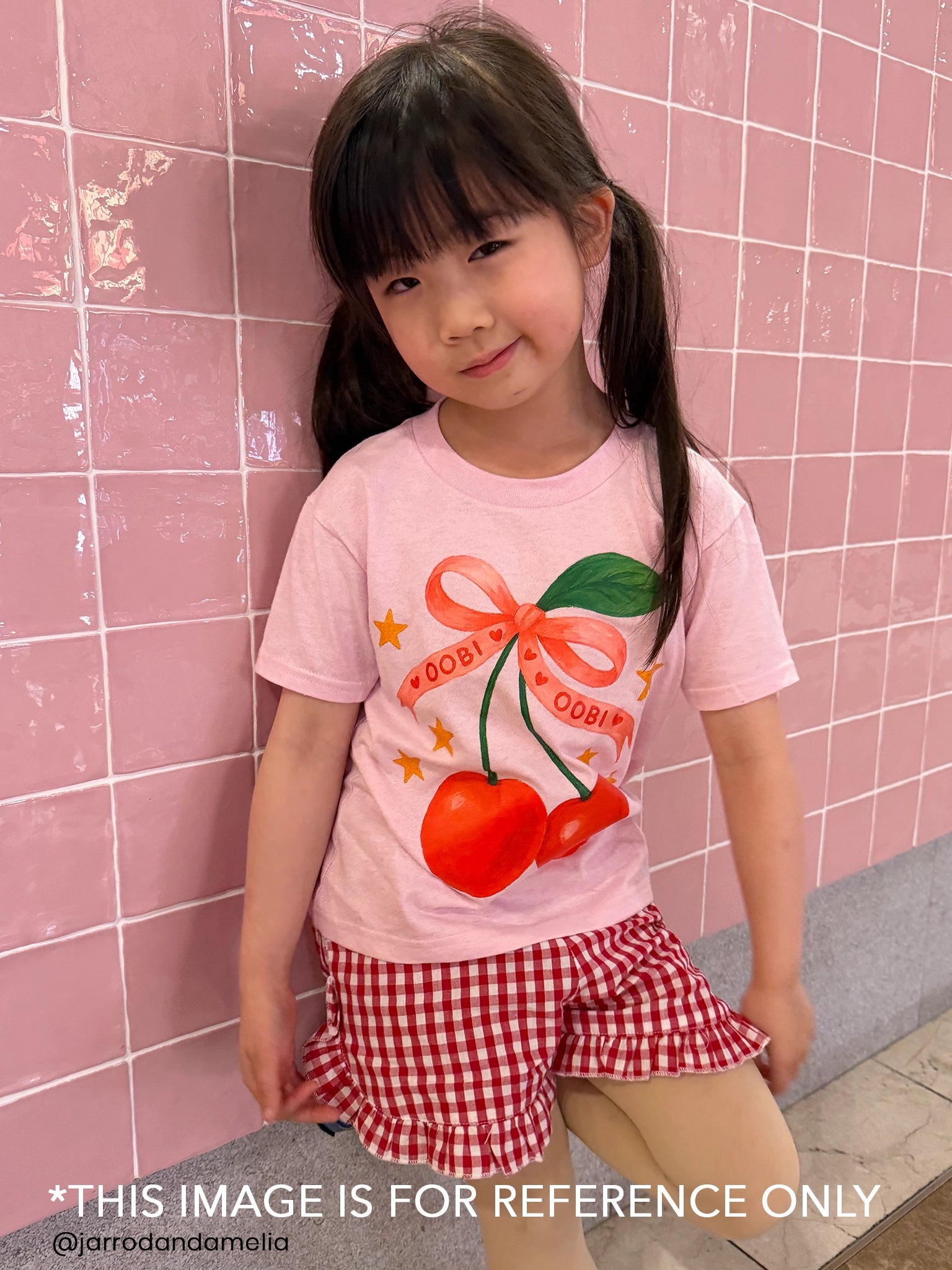 A young girl stands by a pink tiled wall, wearing the Essentials L'amour Printed Tee—ethically made with free personalisation—and red checkered shorts.