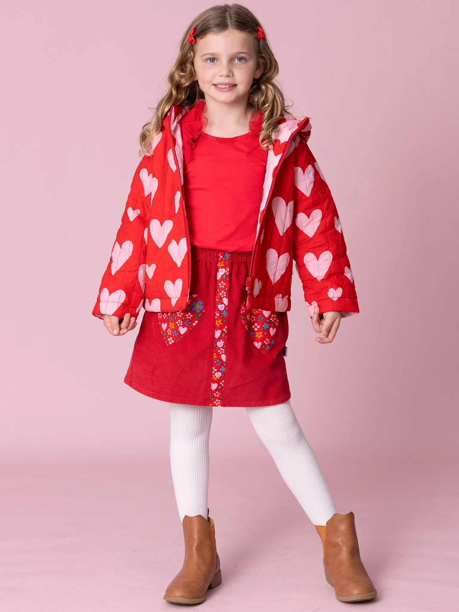Young girl wearing a red heart-patterned jacket and dress on a pink background