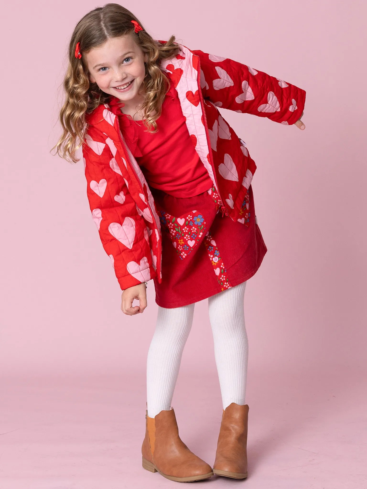 A smiling young girl models the Oobi Raspberry Red Bundle & Save—a heart-print jacket, elastic waist skirt, white tights, and brown boots—posing against a pink background for an adorable look perfect for any occasion.