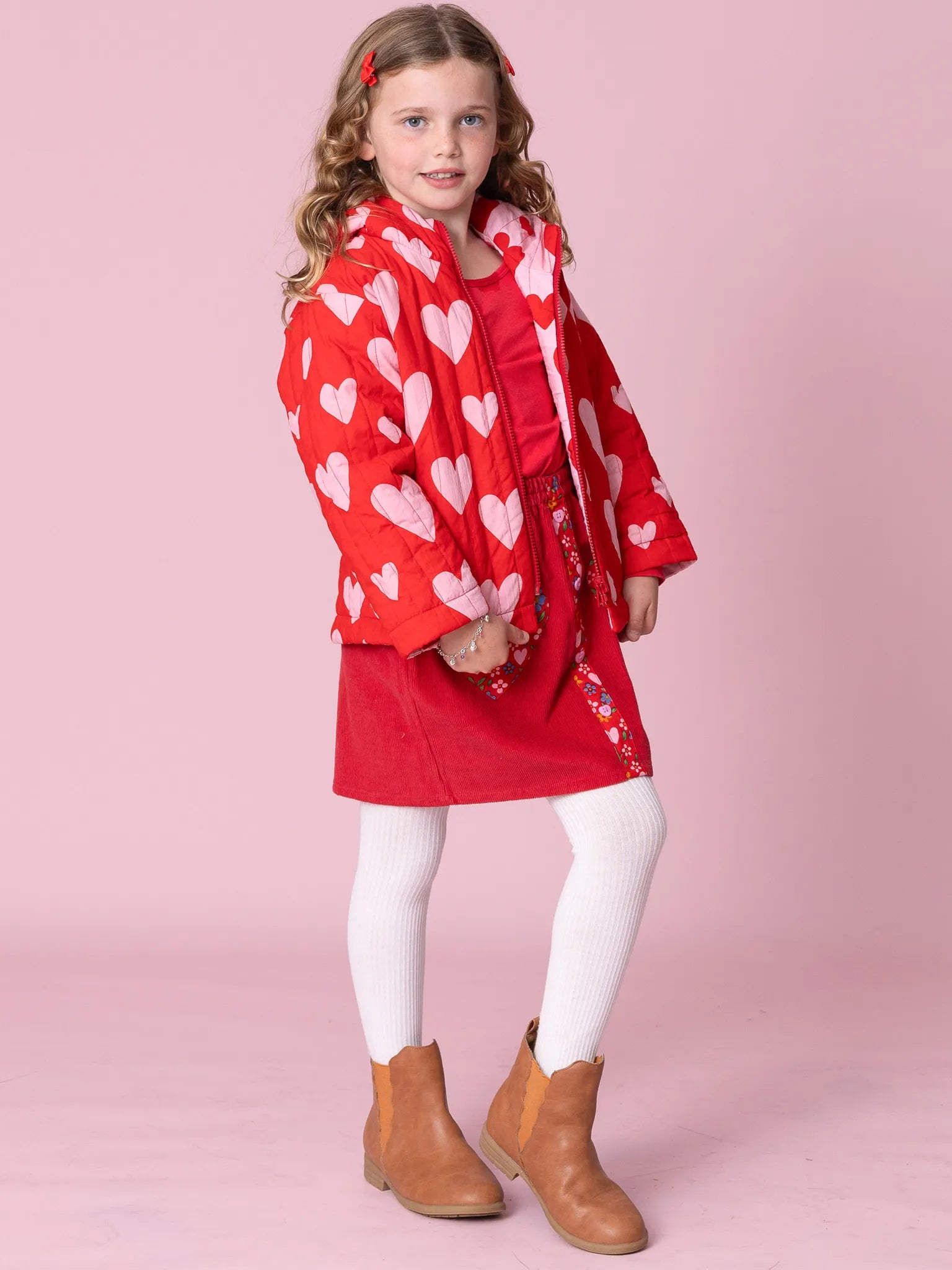 Young girl wearing a red dress with white heart patterns, white tights, and brown boots on a pink background.