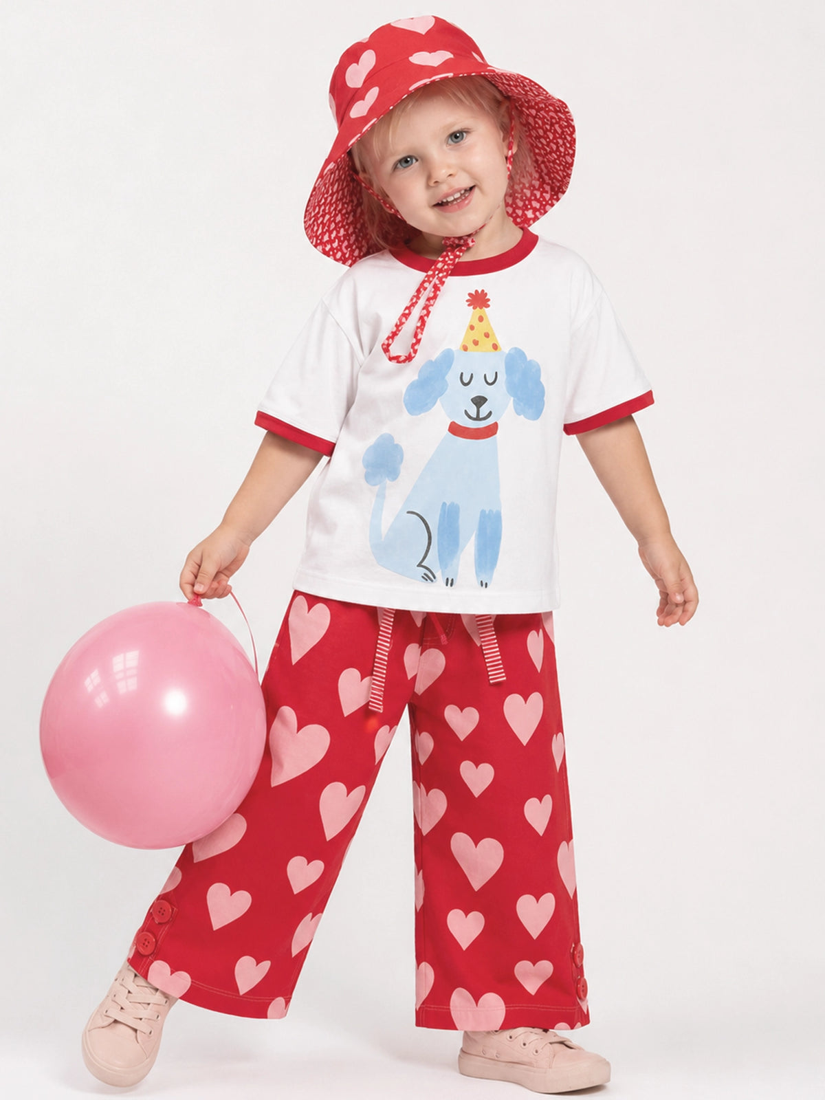 A toddler wearing a matching red and pink children's outfit, including a white ruffle-sleeve shirt with a blue dog graphic, red pants with pink hearts, a red sun hat, and holding a pink balloon against a light gray studio background.