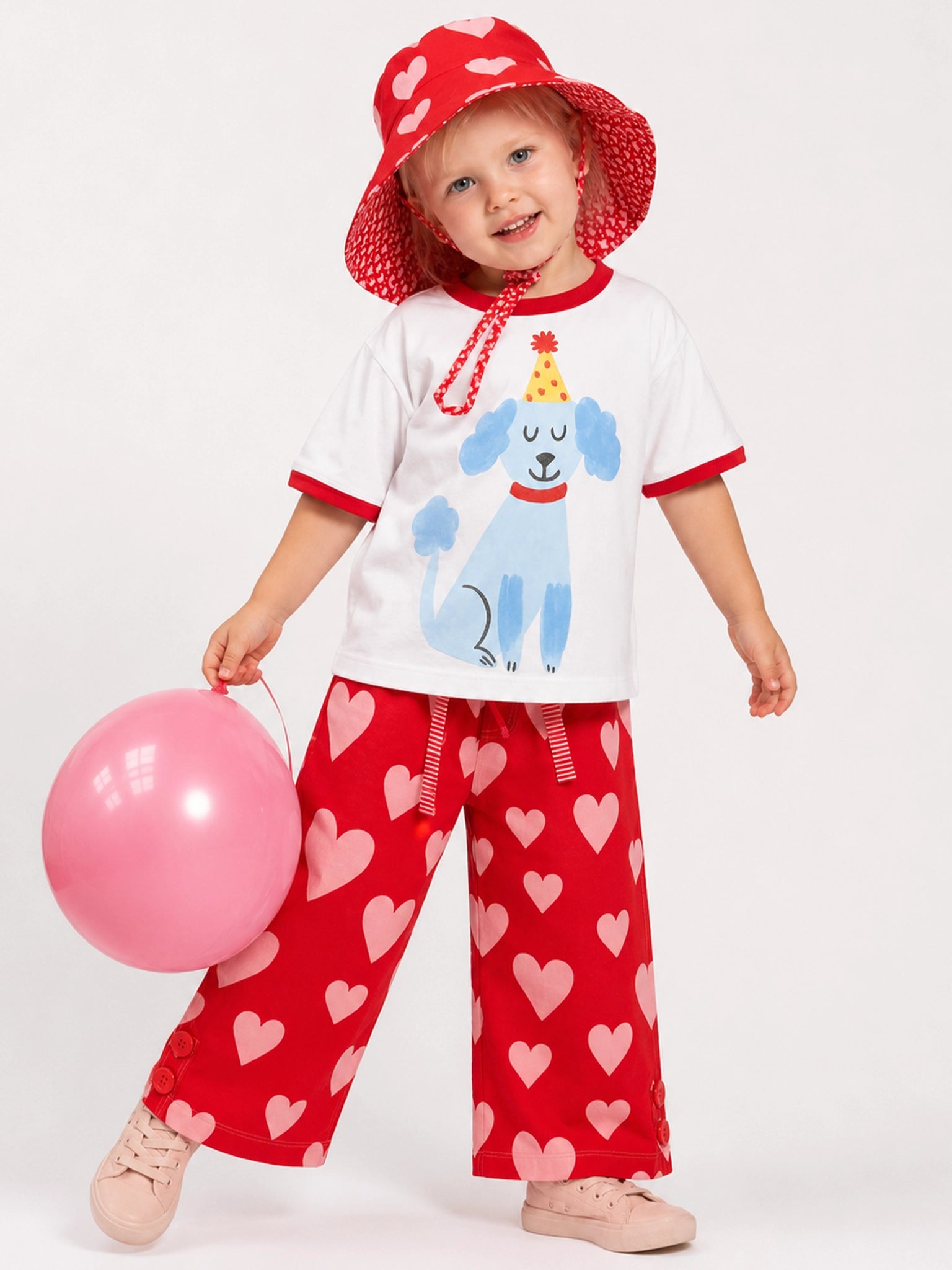 A toddler wearing a matching red and pink children's outfit, including a white ruffle-sleeve shirt with a blue dog graphic, red pants with pink hearts, a red sun hat, and holding a pink balloon against a light gray studio background.