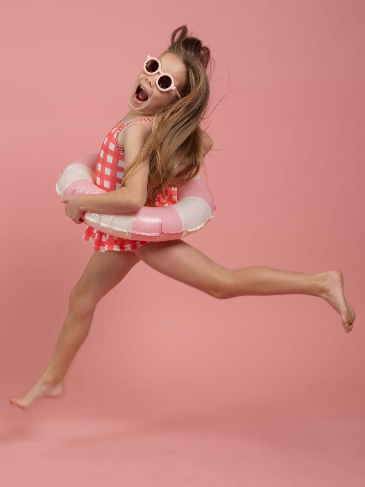 A young girl in Red Gingham’s SPF 50 Gingham Swimsuit with Adjusters and sunglasses jumps mid-air with a pink and white swim ring against a pink background, highlighting playful girls’ swimwear.