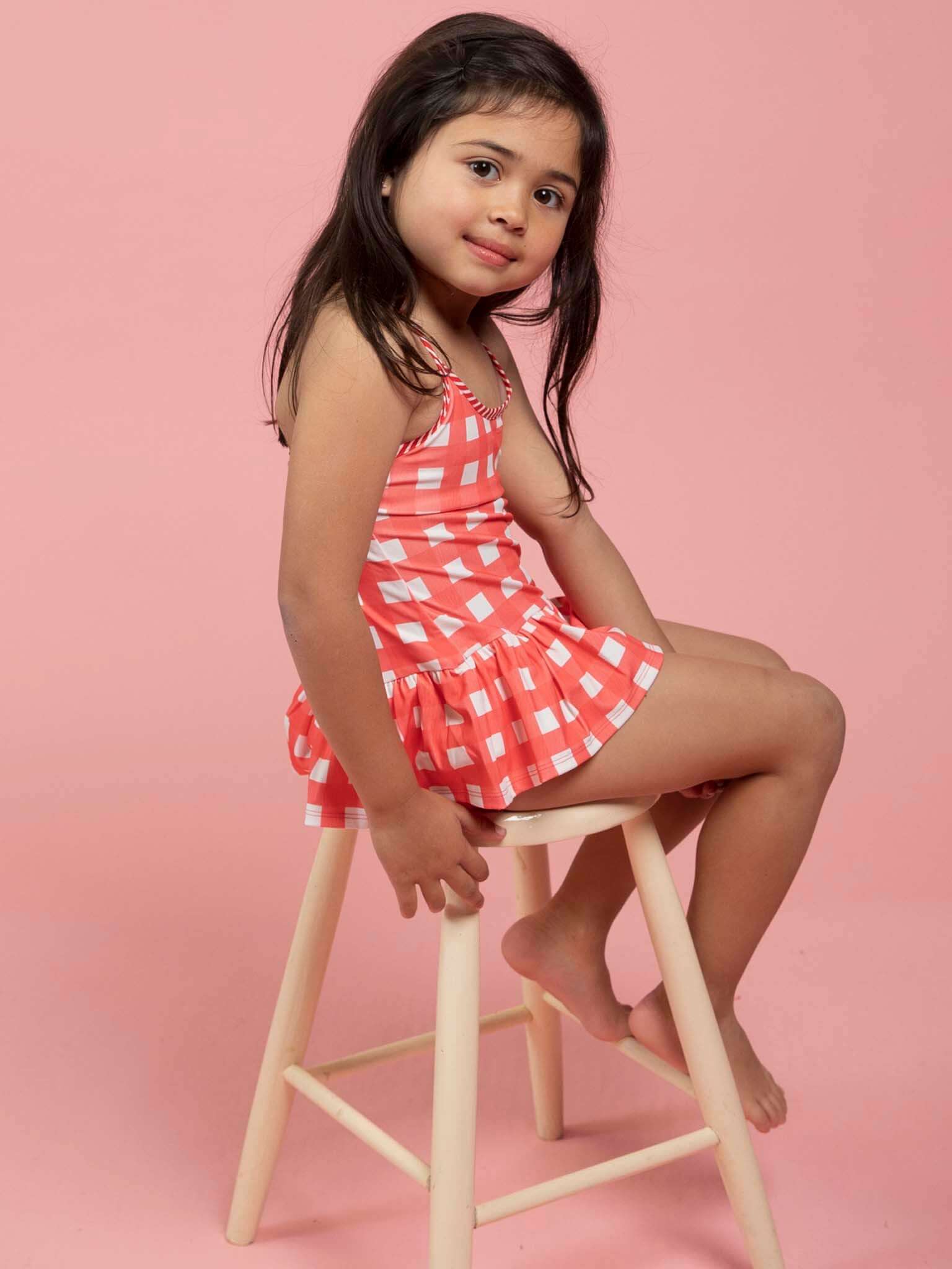 A young girl with long dark hair wears the Red Gingham SPF 50 Gingham Swimsuit with Adjusters in pink and white, sitting sideways on a wooden stool against a pink background.