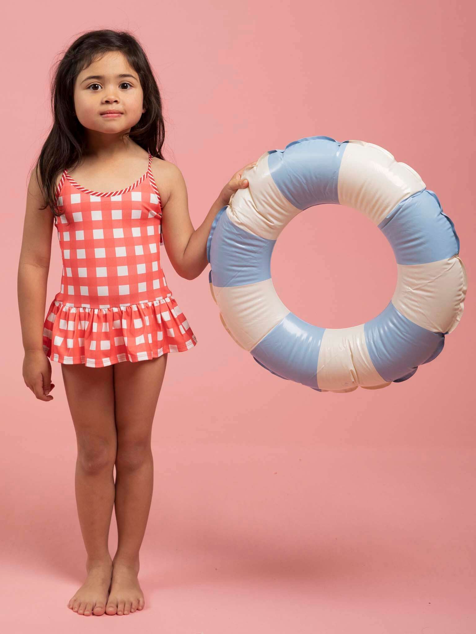 A young girl wearing the Red Gingham SPF 50 Gingham Swimsuit with Adjusters stands barefoot on a pink background, holding a blue and white inflatable swim ring.