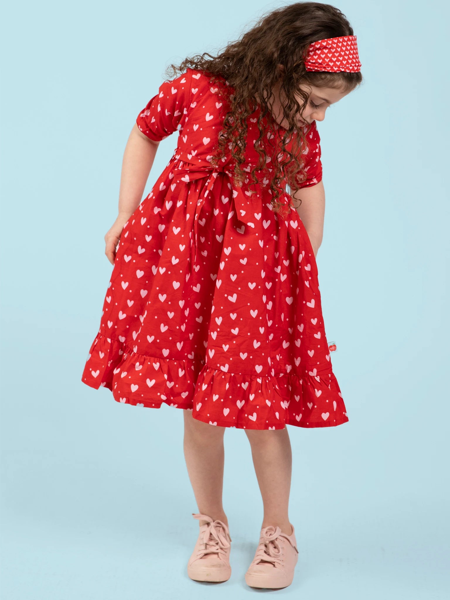 A young girl with curly hair wears the Hearts Red & Pink Heart Midi Dress and a matching headband, looking down against a light blue background.