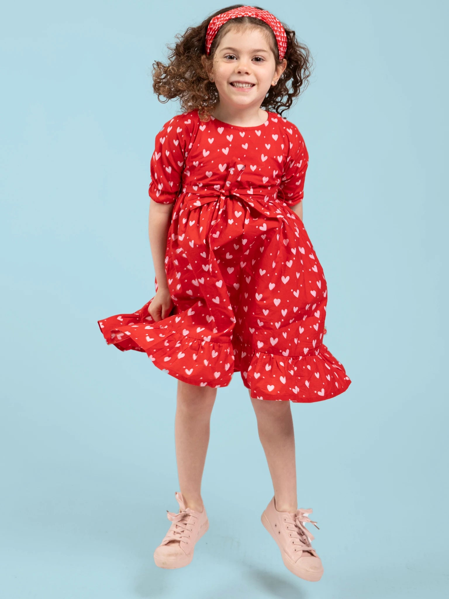 A smiling young girl with curly hair wears the Hearts Red & Pink Heart Midi Dress, made from ethically sourced cotton, paired with a matching headband and pink shoes, jumping against a light blue background.