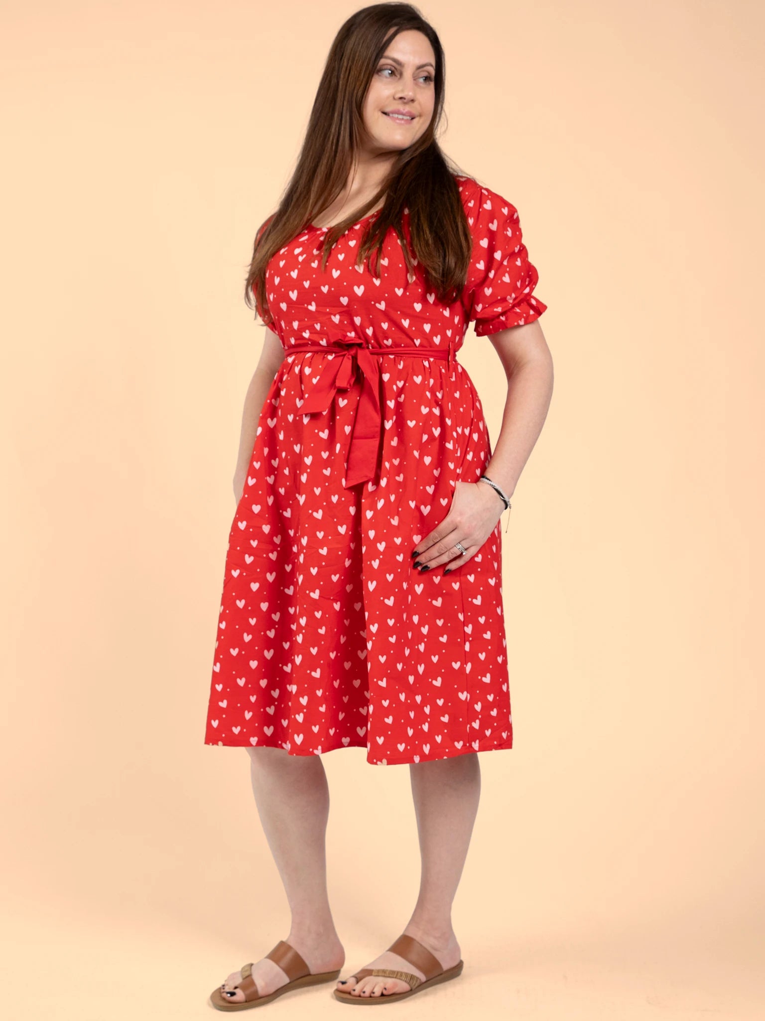 A woman poses against a beige backdrop in the Hearts Pink & Red Heart Women's Midi Dress, featuring hand-printed cotton with a small white heart pattern, waist tie, and paired with brown flat sandals.