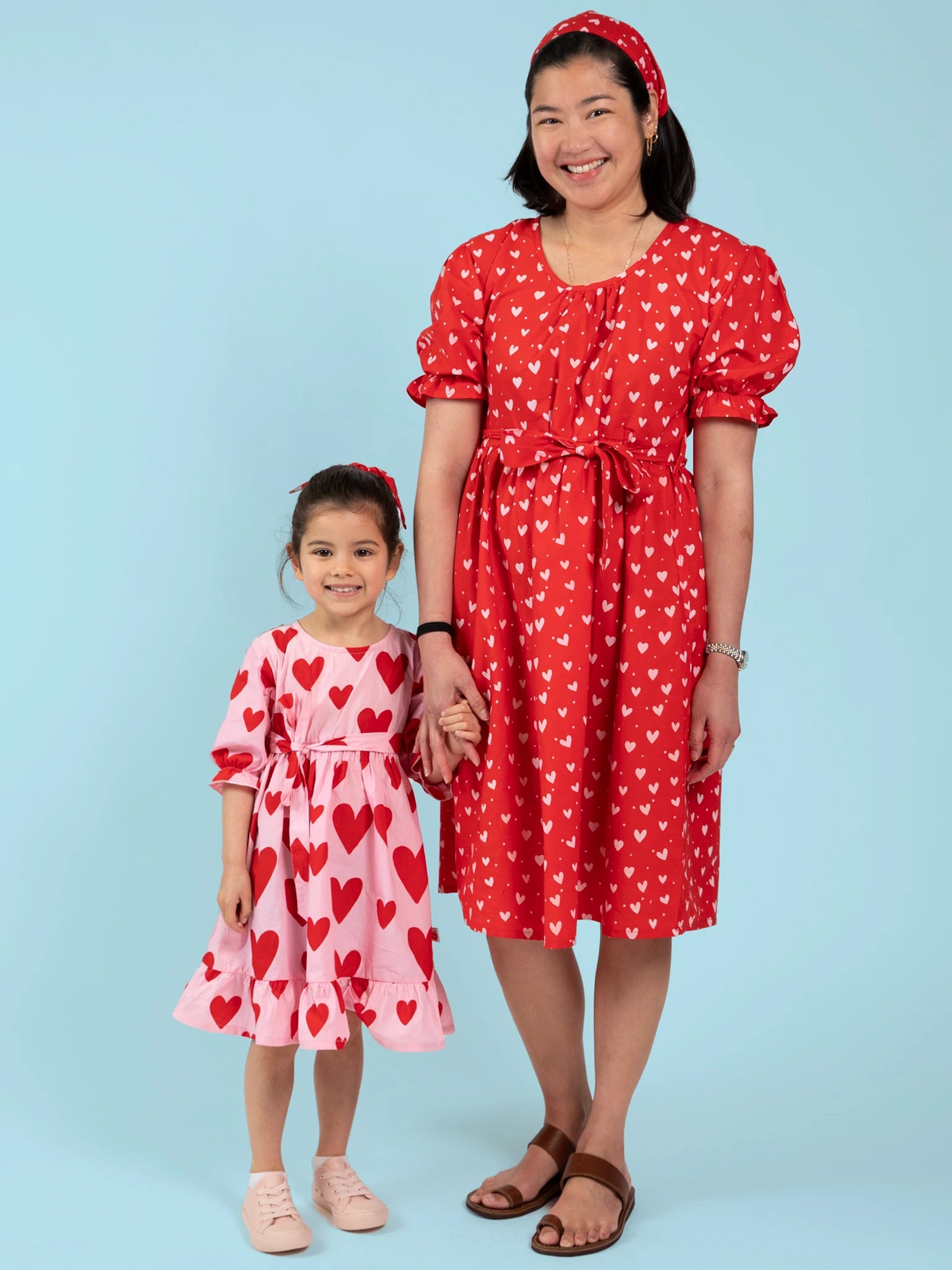 A woman in the Hearts Pink & Red Heart Women's Midi Dress stands side by side and holds hands with a child, both dressed in hand-printed heart patterns against a light blue background.