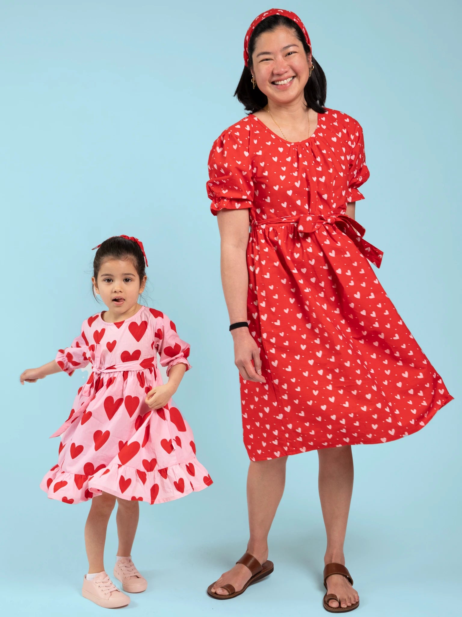 An adult and child stand together in handprinted cotton dresses with red hearts: the adult wears the Big Hearts midi dress in red, while the child wears the Big Heart Girl's Midi Dress in light pink; both are ethically made in India.