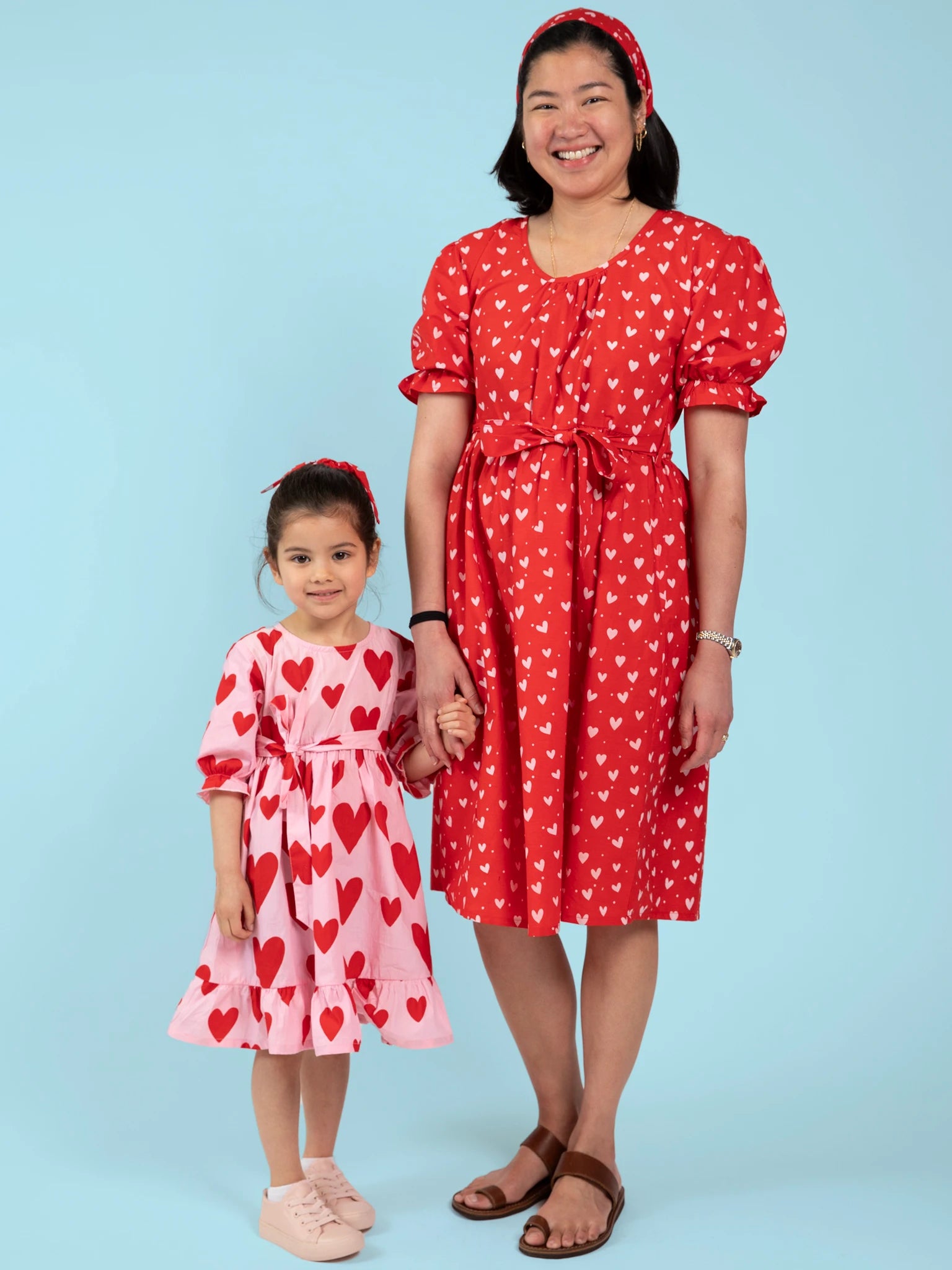 An adult and child hold hands wearing handprinted cotton dresses with heart patterns; the adult in a red Big Heart midi dress, and the child in a pink Big Hearts Big Heart Girl's Midi Dress—both ethically made in India.