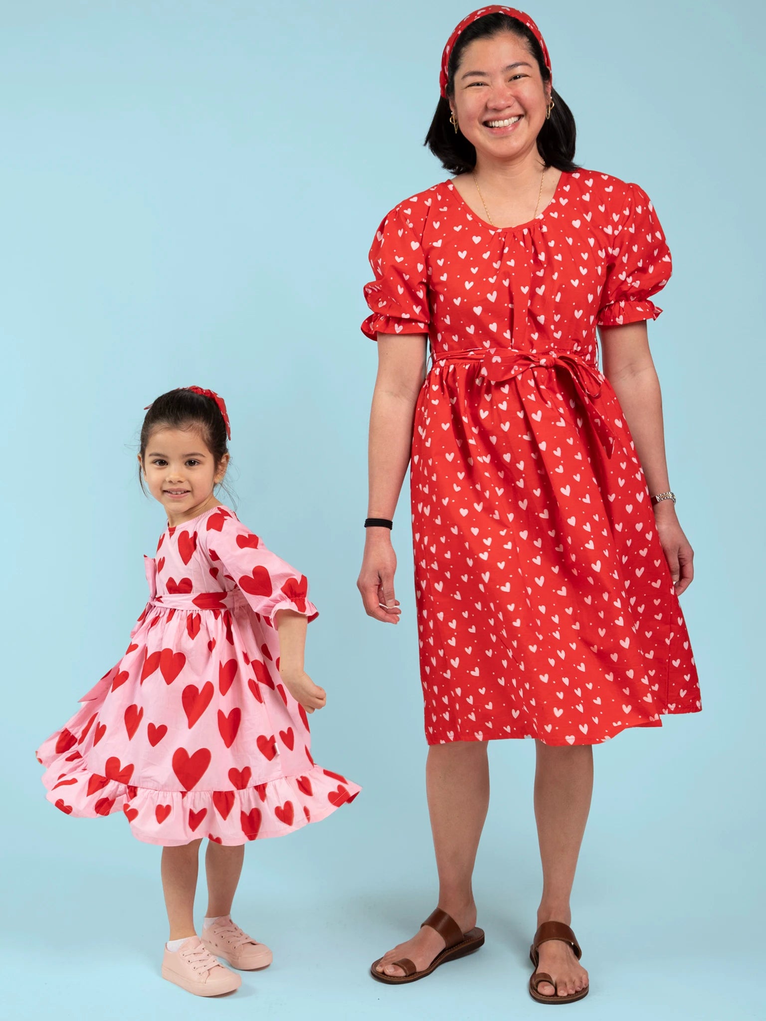 An adult and child stand together wearing Big Hearts’ handprinted cotton Big Heart Girl’s Midi Dresses in red and white heart motifs, set against a light blue background. These charming dresses are ethically made in India.