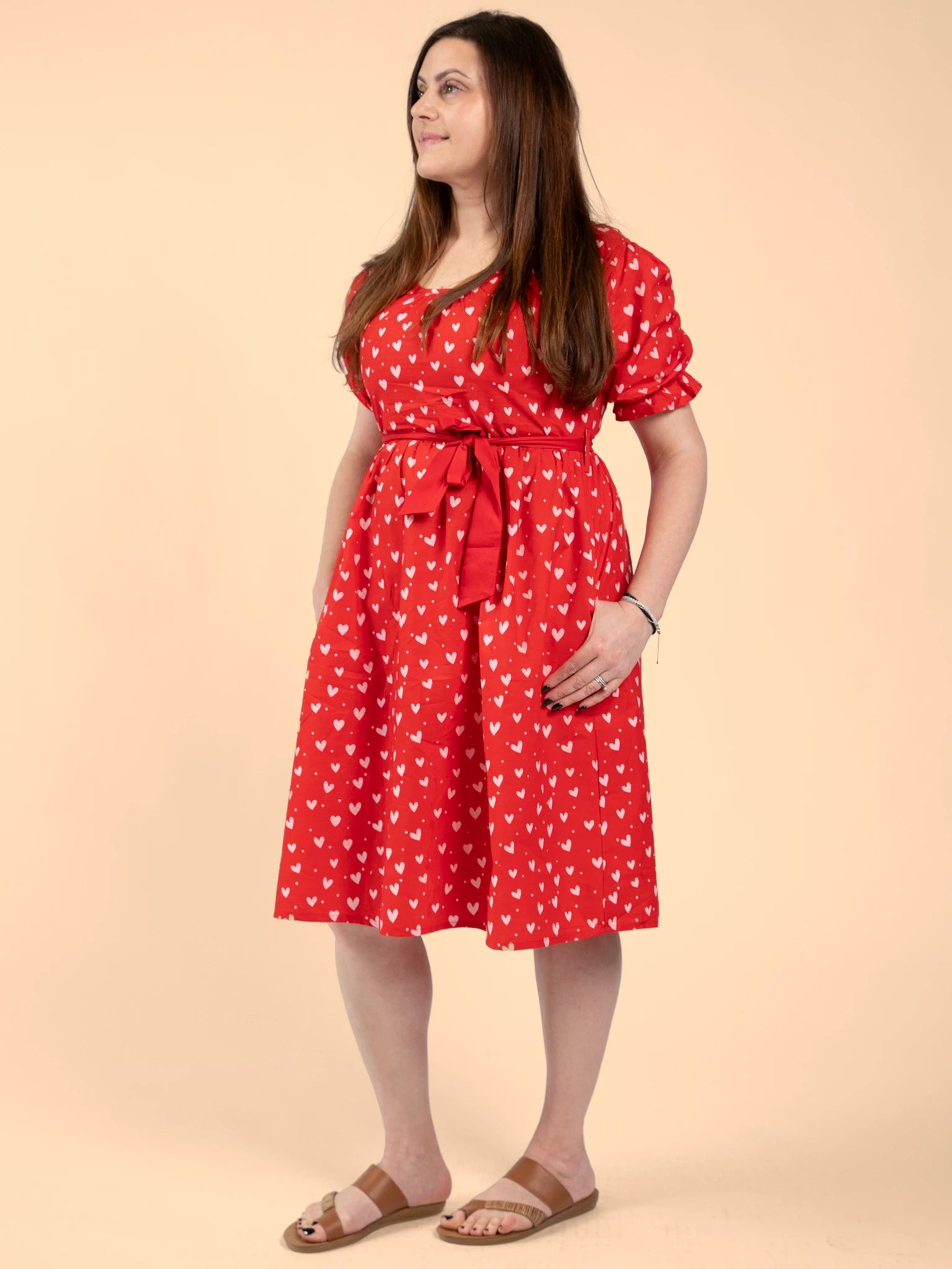 A woman poses against a beige background in the Hearts Pink & Red Heart Women's Midi Dress, featuring an ethical hand-printed design with white heart patterns. She completes the look with sandals and a wristwatch.