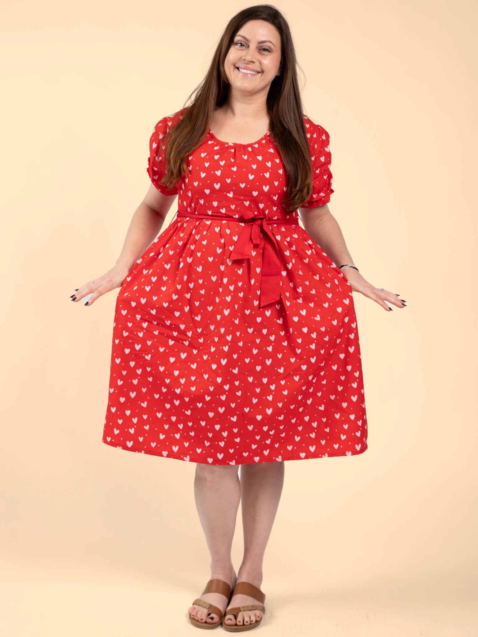 A woman stands against a beige background wearing the Hearts Pink & Red Heart Women's Midi Dress featuring a hand-printed heart pattern, short puff sleeves, tied waist, and brown sandals.