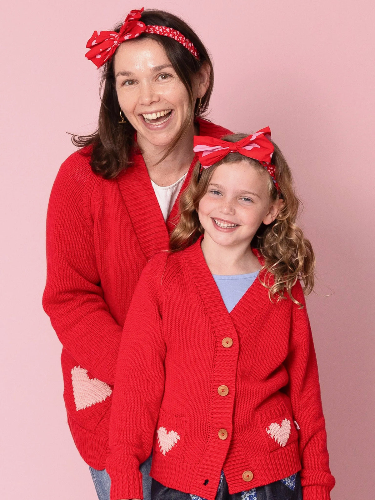 Woman and young girl wearing matching red cardigans with heart designs on a pink background