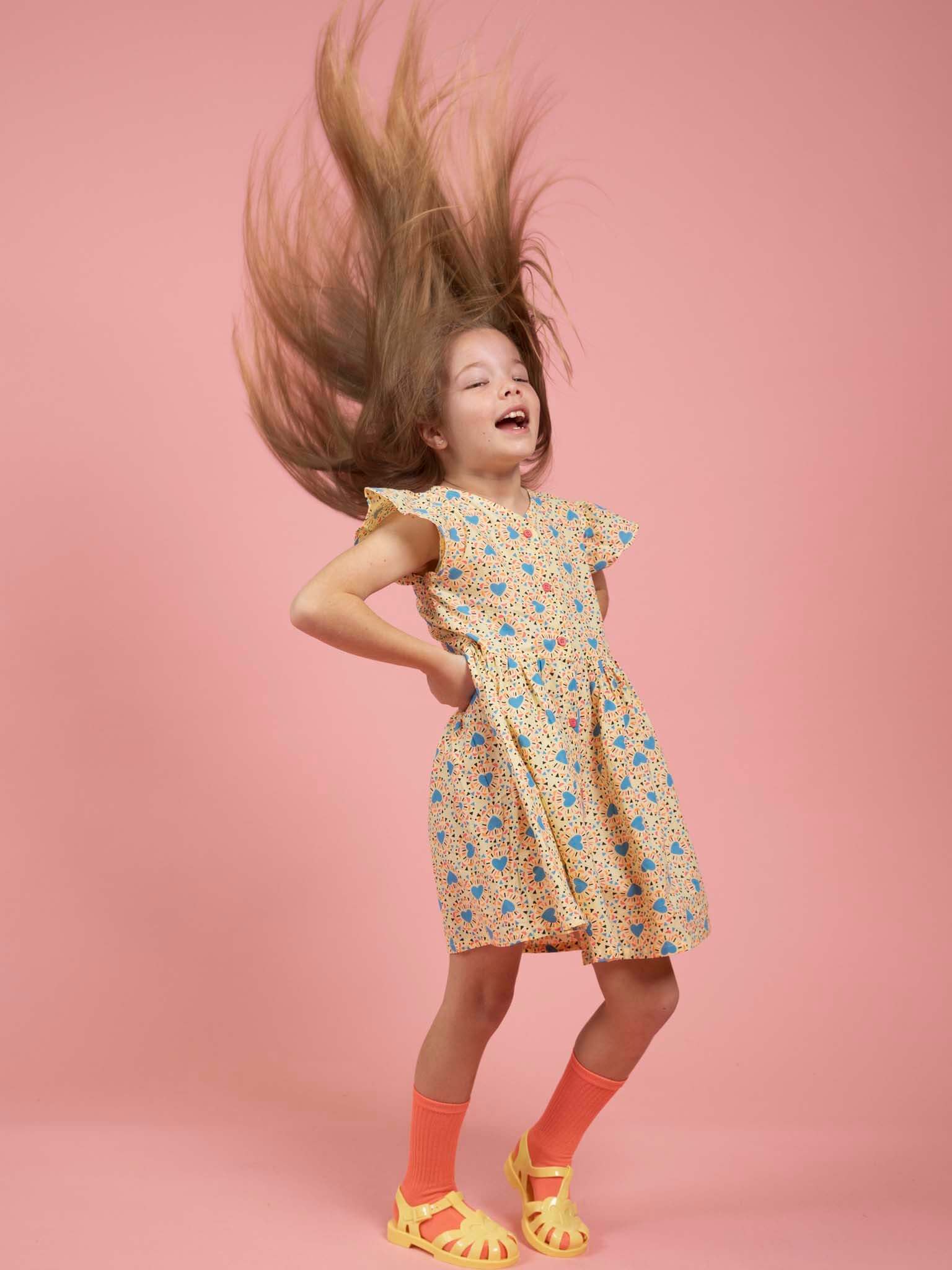 A young girl poses against a pink background with her hair flying upwards, wearing the Starburst Astra Dress by Starburst—featuring cap sleeves and machine-washable cotton—styled with orange socks and yellow sandals.