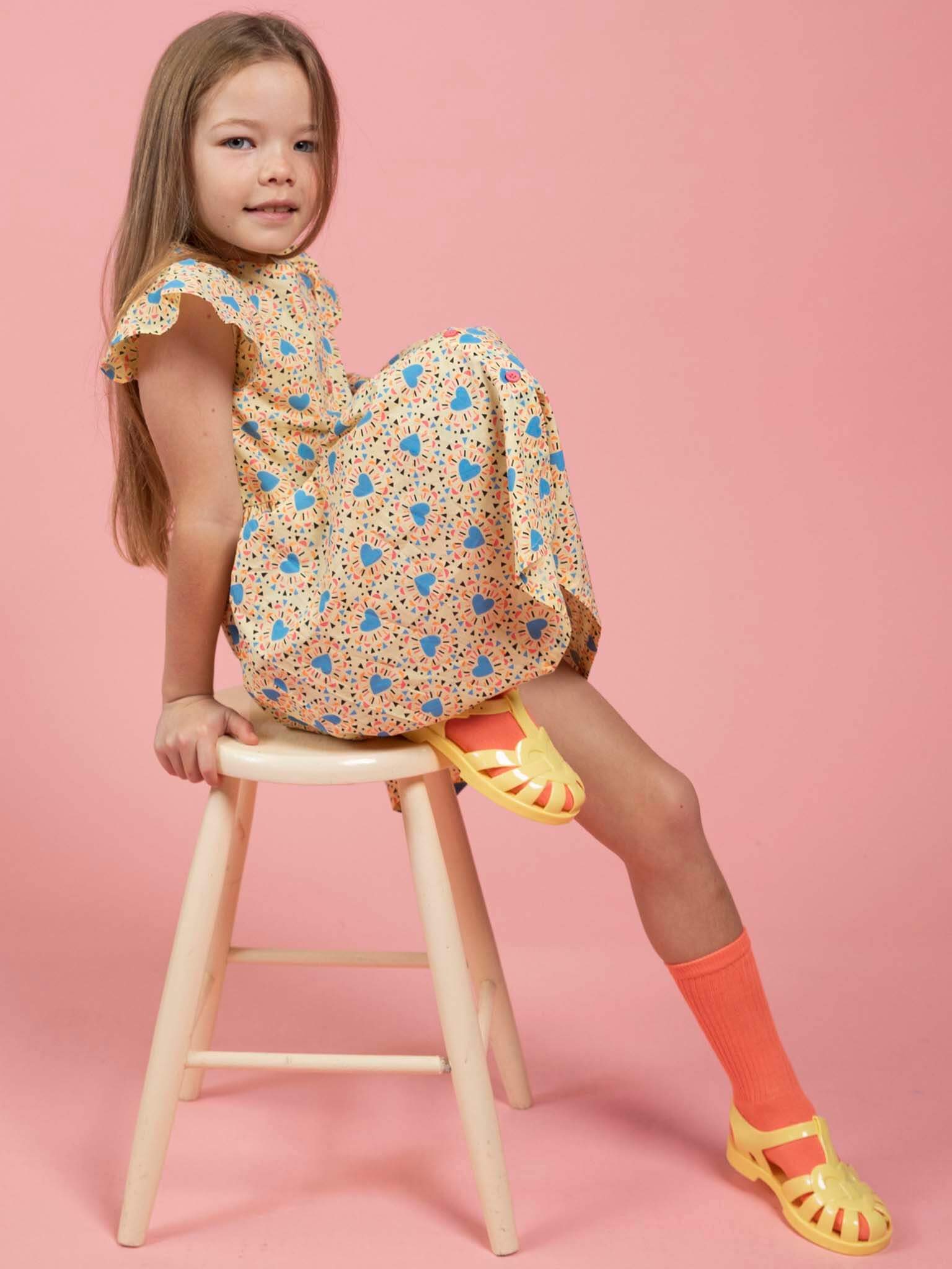 A young girl with long hair sits sideways on a wooden stool, wearing the Starburst Astra Dress by Starburst with orange socks and yellow sandals, set against a pink background.