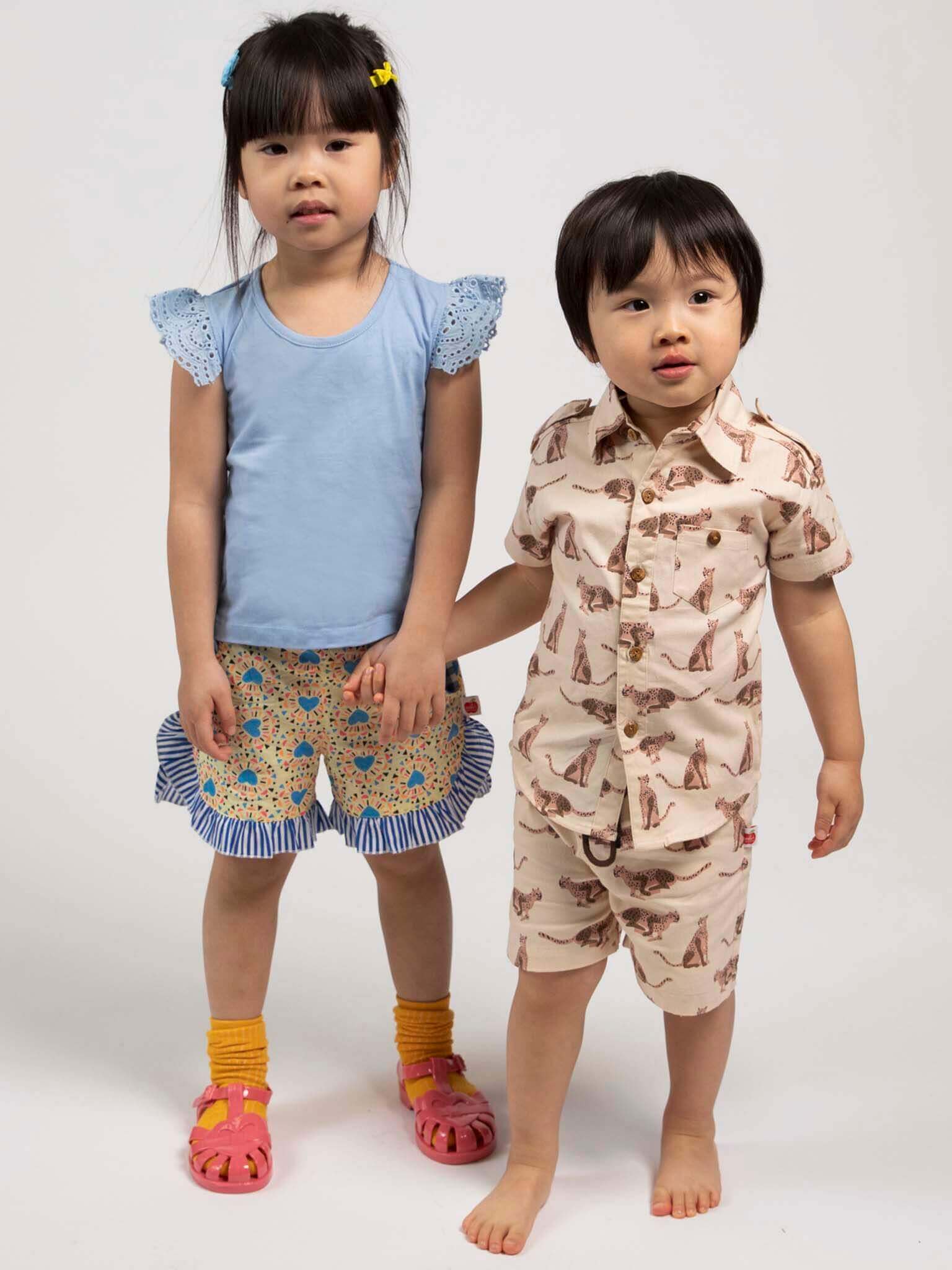 Two children holding hands, wearing matching outfits with ruffled sleeves and shorts on a white background.
