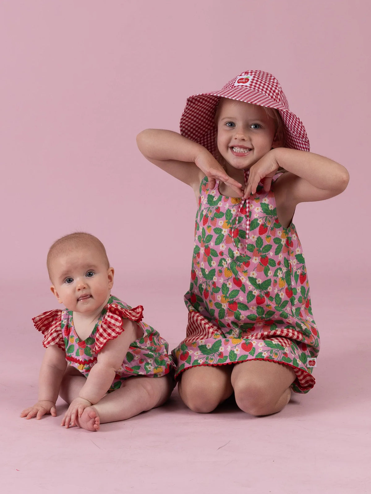 Big sister in the Strawberry Fields Gracie dress and gingham Chloe hat beside baby in the matching romper