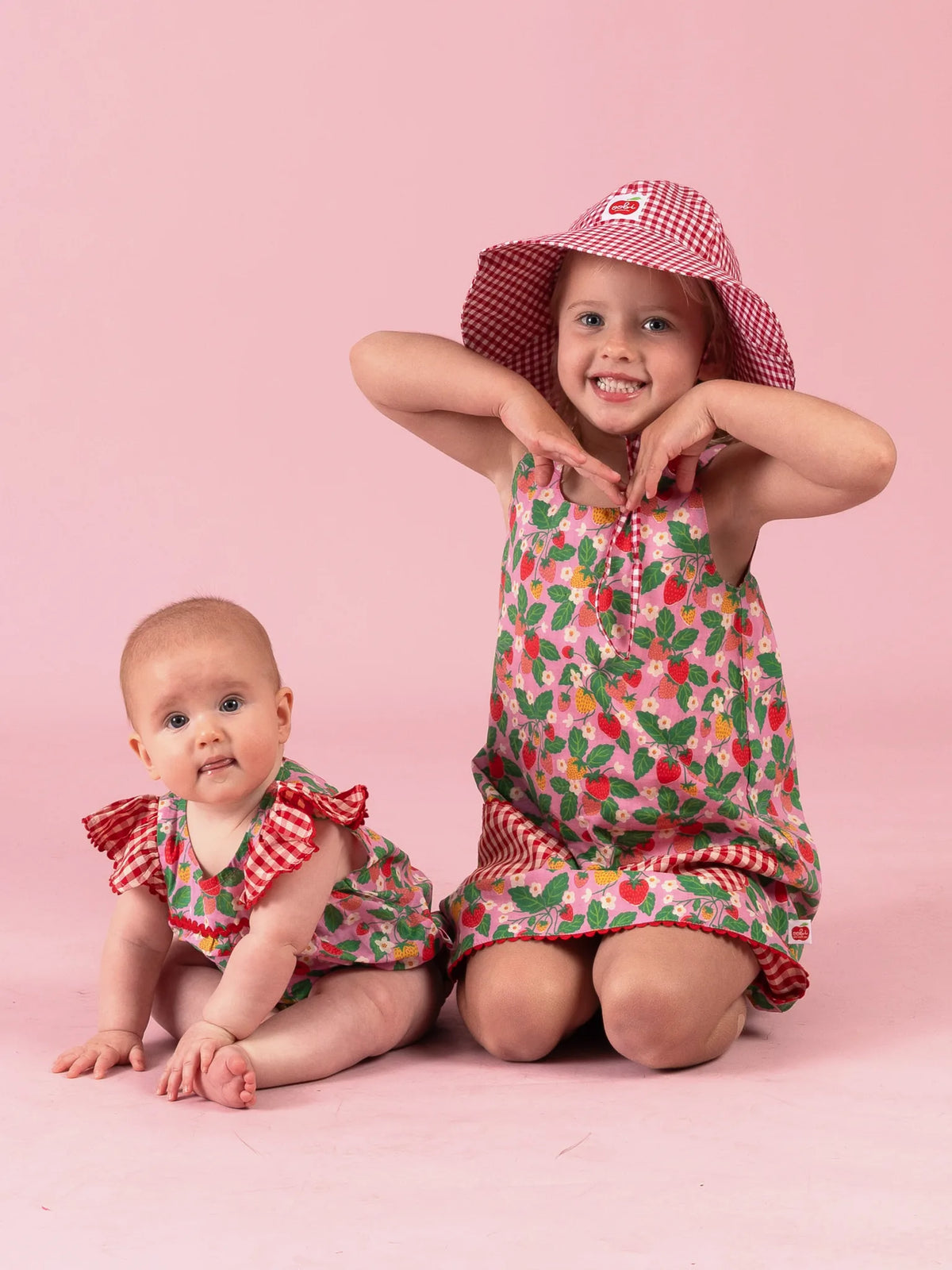 Big sister in the Strawberry Fields Gracie dress and gingham Chloe hat beside baby in the matching romper