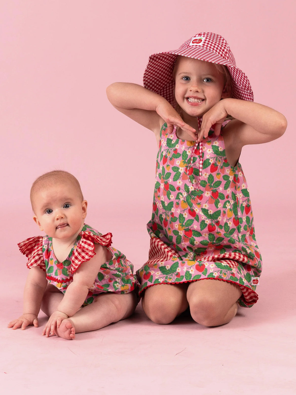 Big sister in the Strawberry Fields Gracie dress and gingham Chloe hat beside baby in the matching romper
