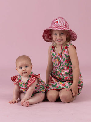 Smiling girl in the Strawberry Fields Gracie dress and gingham Chloe sun hat with her baby sister