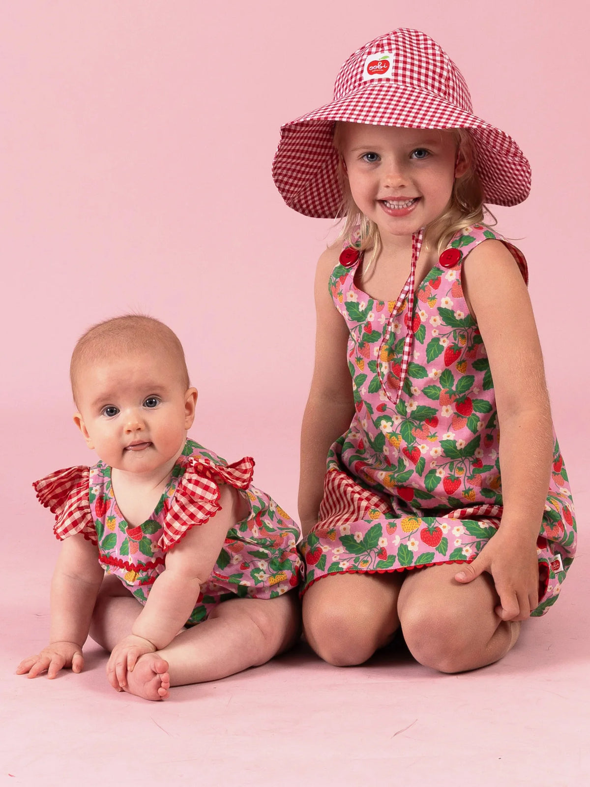 Smiling girl in the Strawberry Fields Gracie dress and gingham Chloe sun hat with her baby sister