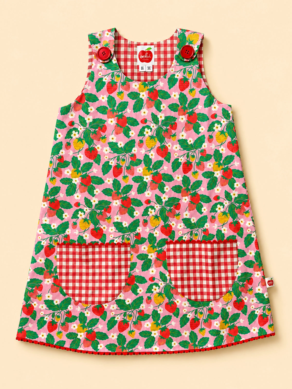 A sleeveless children's dress with a pink strawberry and leaf print, featuring red and white gingham pockets and shoulder straps, displayed on a solid beige background.