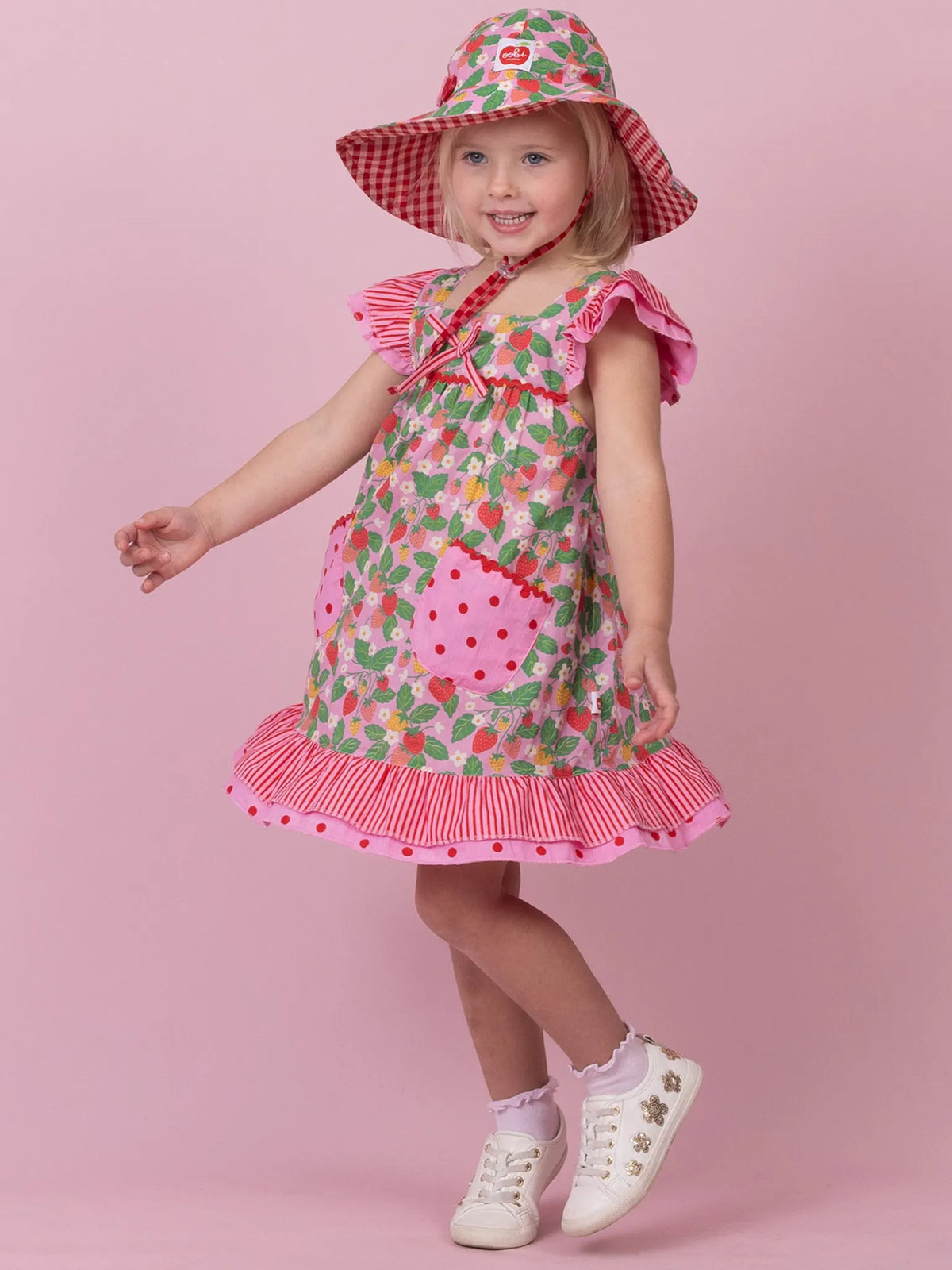 A young girl modeling a strawberry-themed dress, matching sun hat, and gingham accessories against a solid pink studio background.