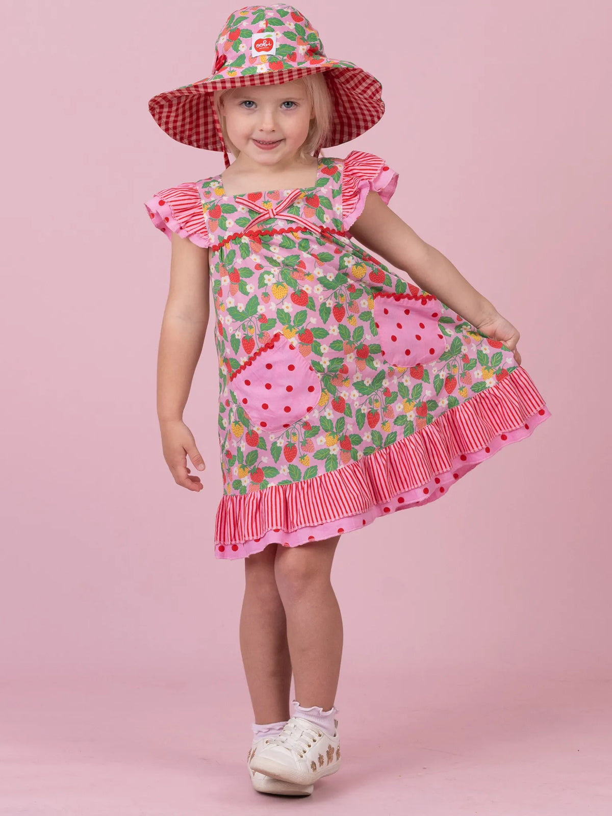 A young girl modeling a strawberry-themed dress and matching sun hat against a solid pink studio background. The dress features a green and red strawberry print with pink ruffled trim and pockets, and she is wearing white shoes.