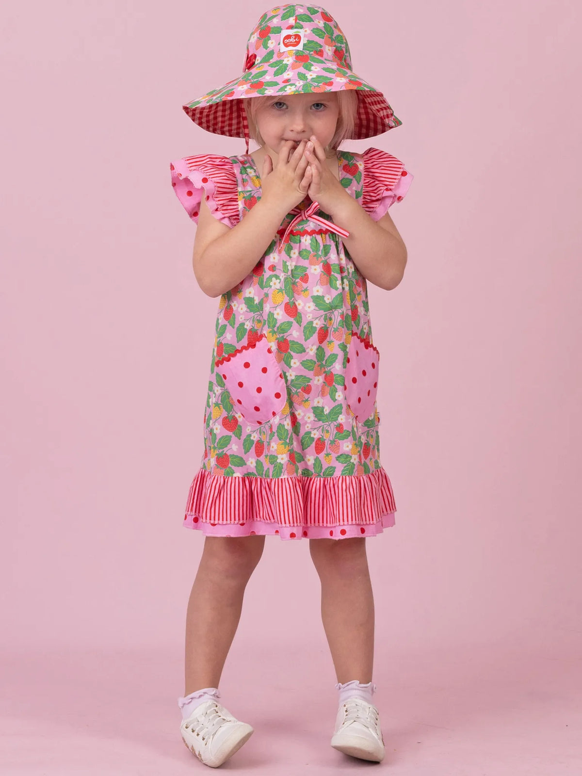 A young child modeling a strawberry-themed dress and matching sun hat against a solid pink studio background. The dress features a green and red strawberry print with pink ruffled trim, and the matching hat has a red and white gingham brim and pink floral crown.