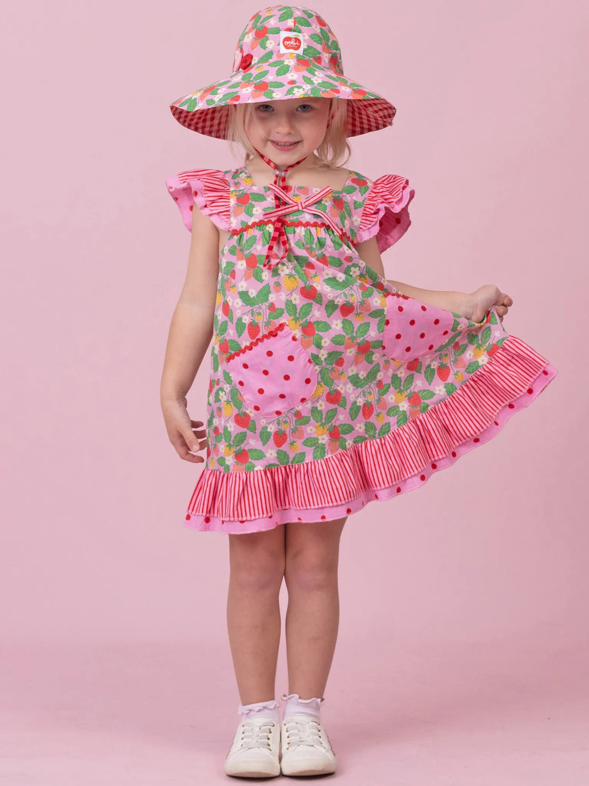 A young girl modeling a strawberry-themed dress and matching sun hat against a solid pink studio background. The dress features a green and red strawberry print with pink ruffled trim, and the matching hat has a red and white gingham pattern with a strawberry design.