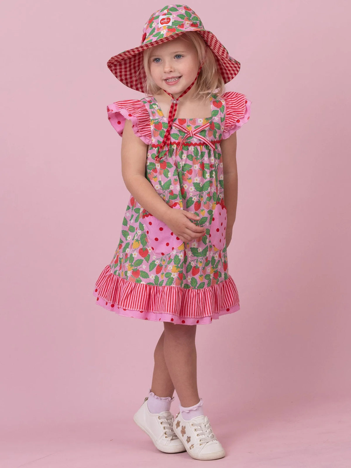 A young girl modeling a strawberry-themed dress and matching sun hat against a solid pink studio background. The dress features a pink and white gingham striped hem, ruffled sleeves, and a watermelon print with pockets, while the hat has a matching gingham and strawberry pattern.