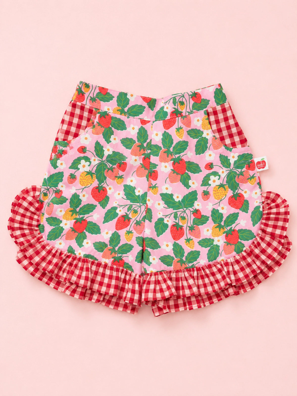 Pink shorts with a strawberry and leaf print, featuring red and white gingham ruffle trim at the bottom and side pockets, laid flat on a solid pink background.