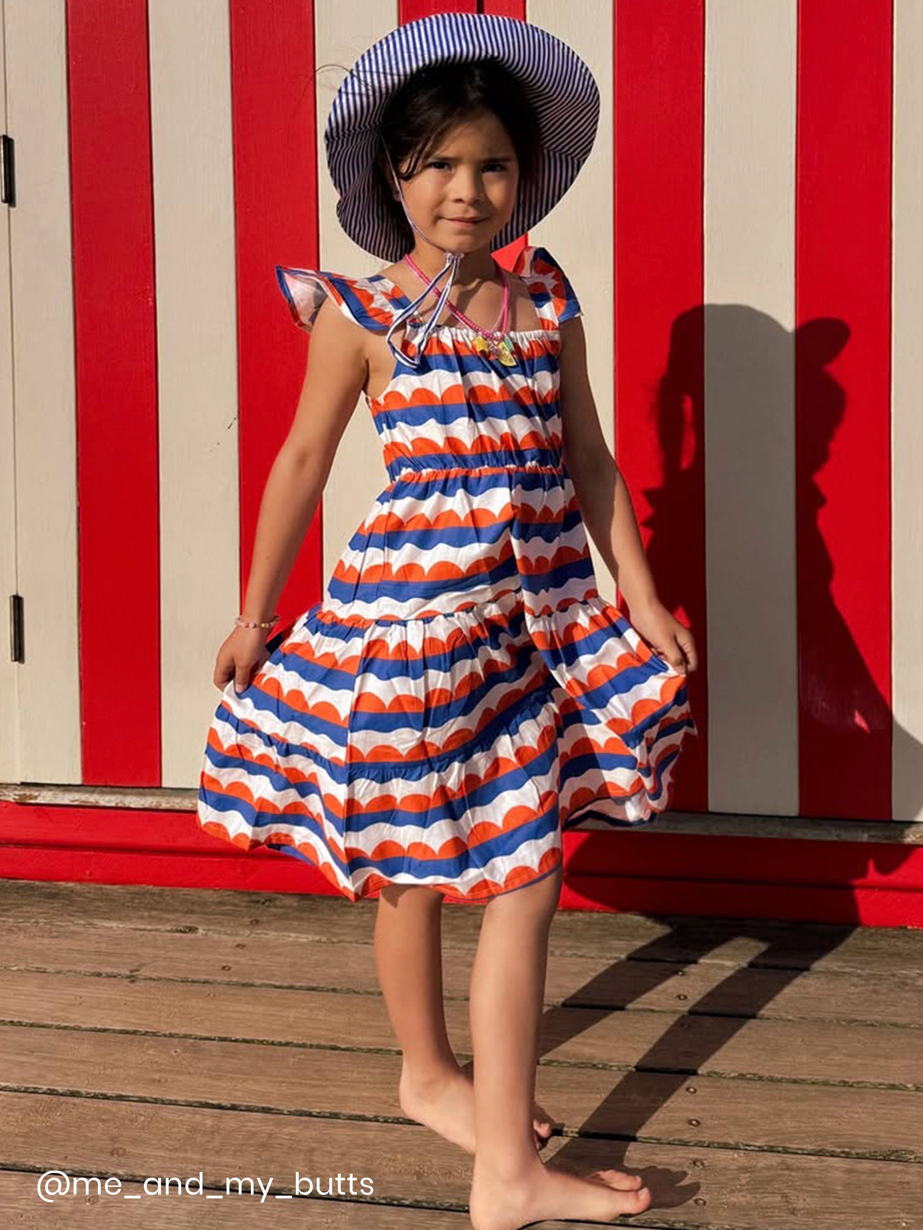 A young girl in the Nautical Holiday Dress by Nautical and a summer hat stands barefoot on a wooden deck before a red and white striped wall.