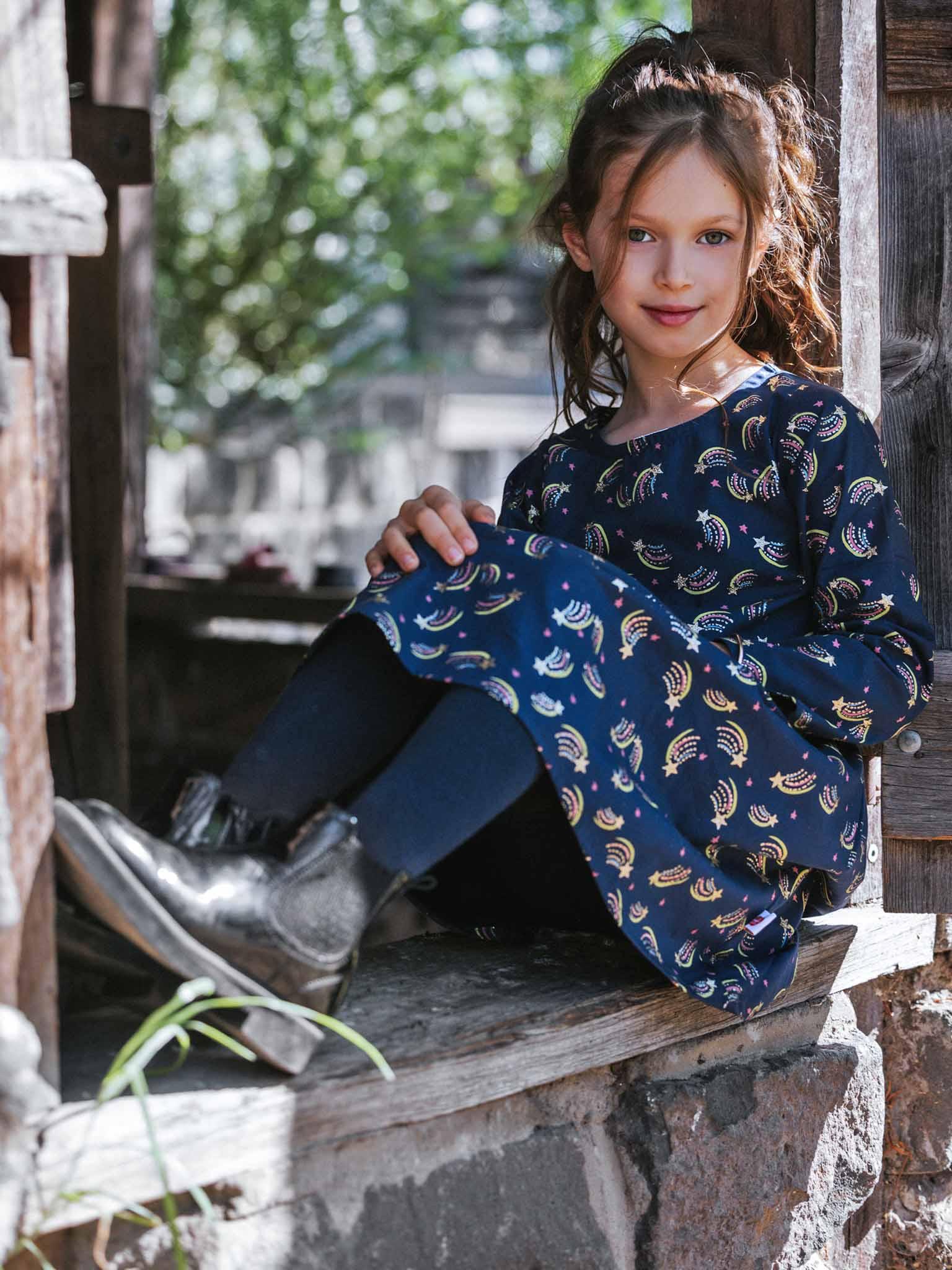 A young girl wears the Oobi Superstar Dress, featuring a sparkly star design, and silver boots as she sits on an outdoor wooden windowsill, looking at the camera.