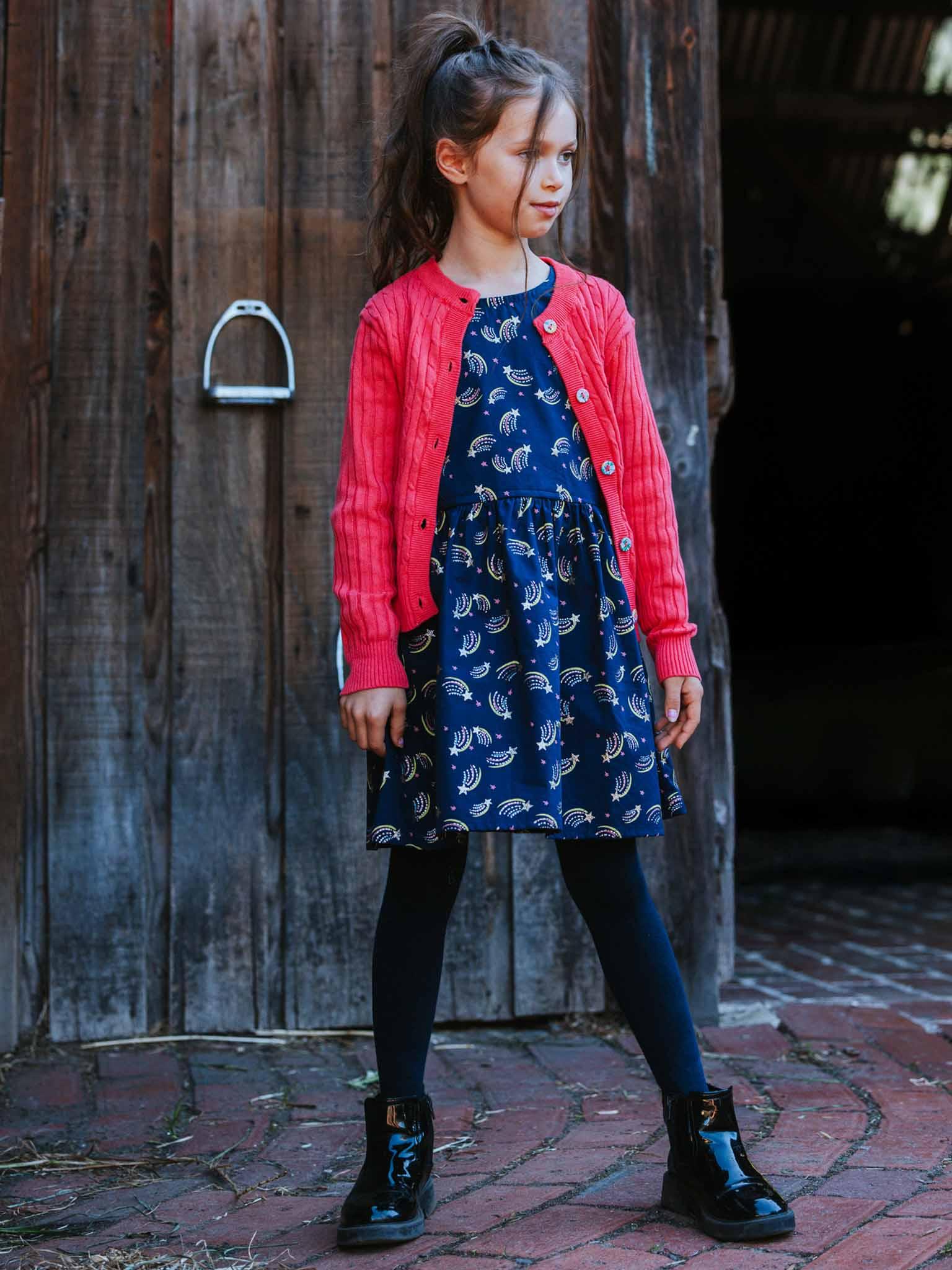 A young girl wears the Oobi Superstar Dress with a sparkly star design, paired with a red cardigan, black leggings, and black boots, standing on bricks in front of a wooden barn door.