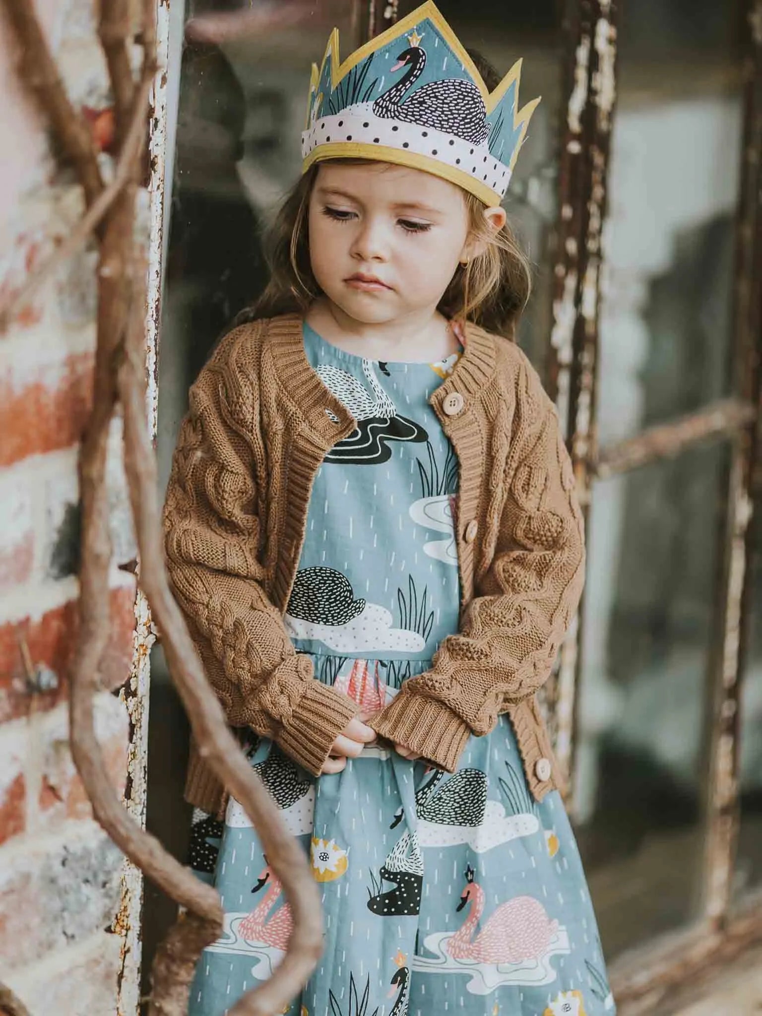 Child wearing a patterned crown and brown cardigan in front of a textured wall.
